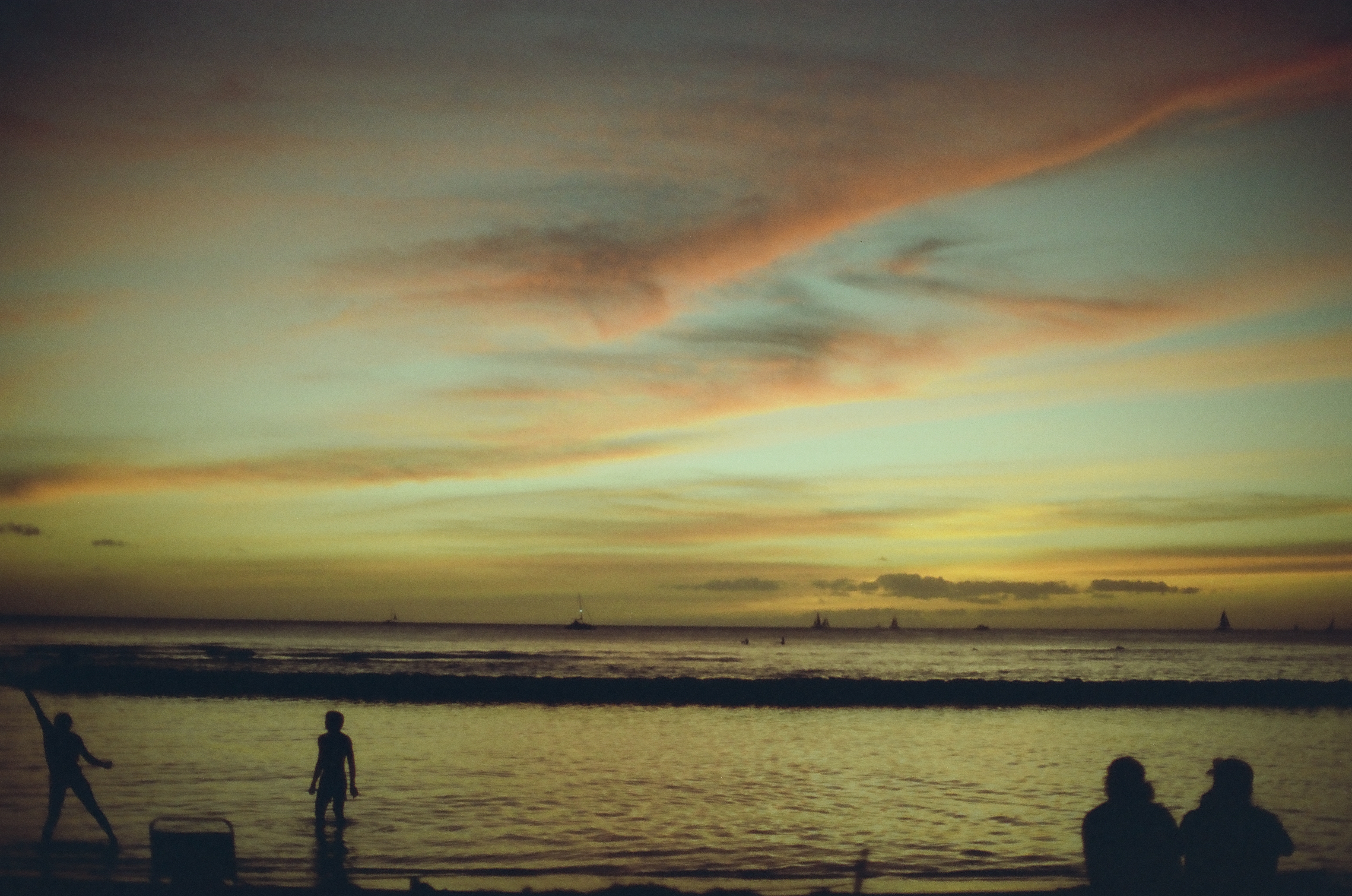 Small figures stand beside calm water beneath long pink clouds and a pale green sky.