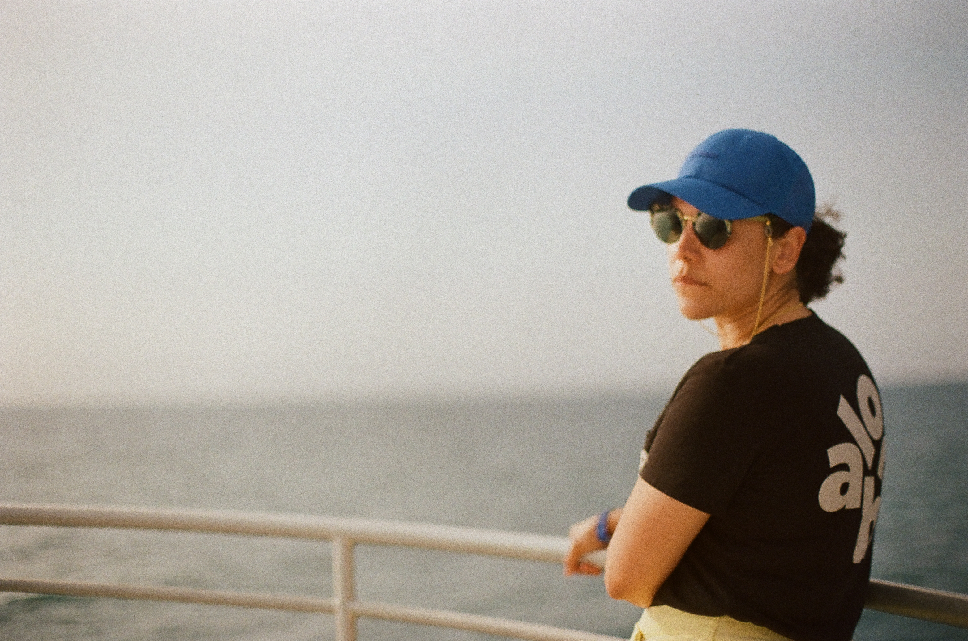 A person in a blue cap and dark shirt stands in profile beside a railing with calm water behind.
