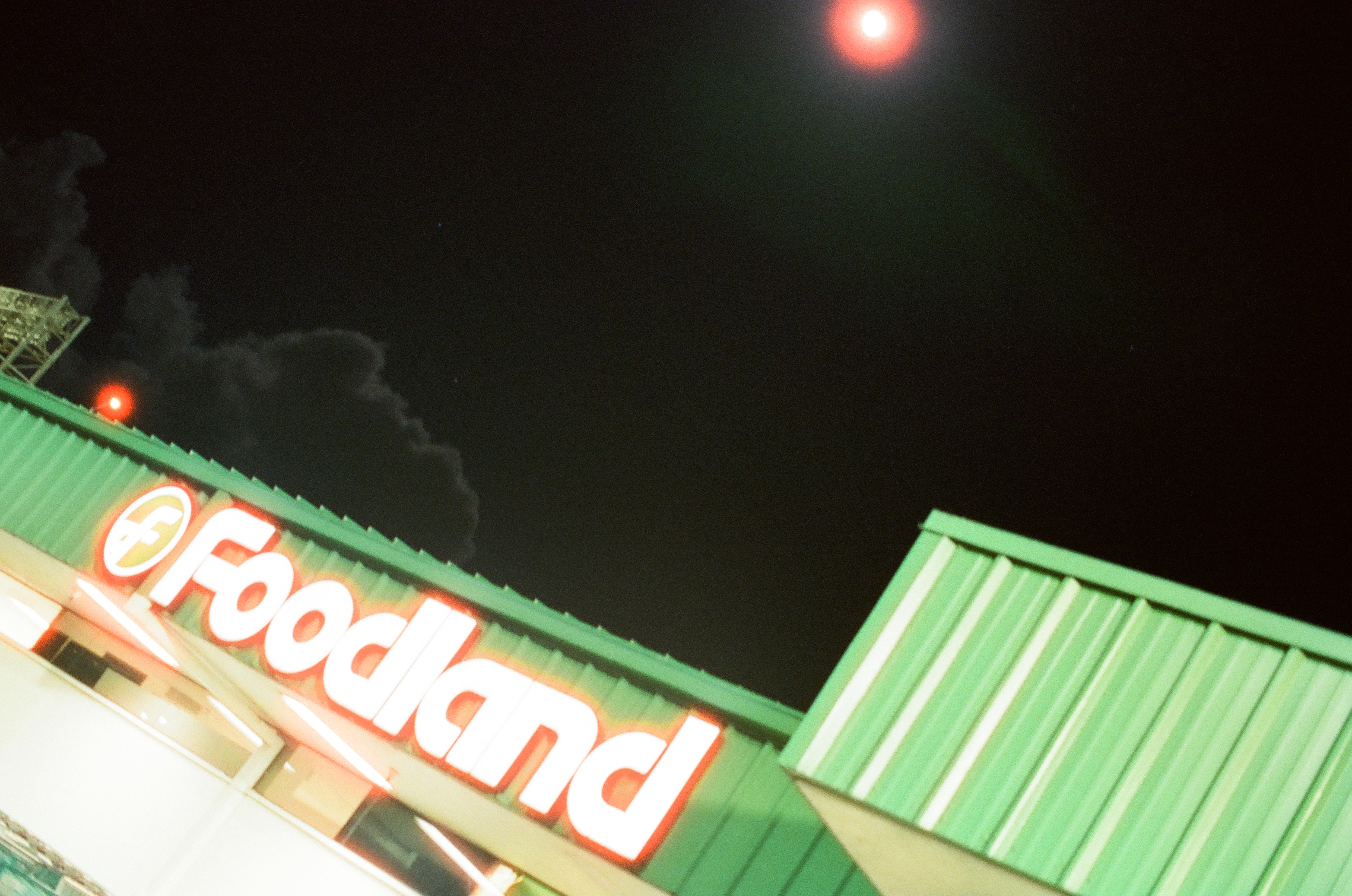 A glowing red food sign sits on an angled green roof beneath a black sky and tall cloud plume.