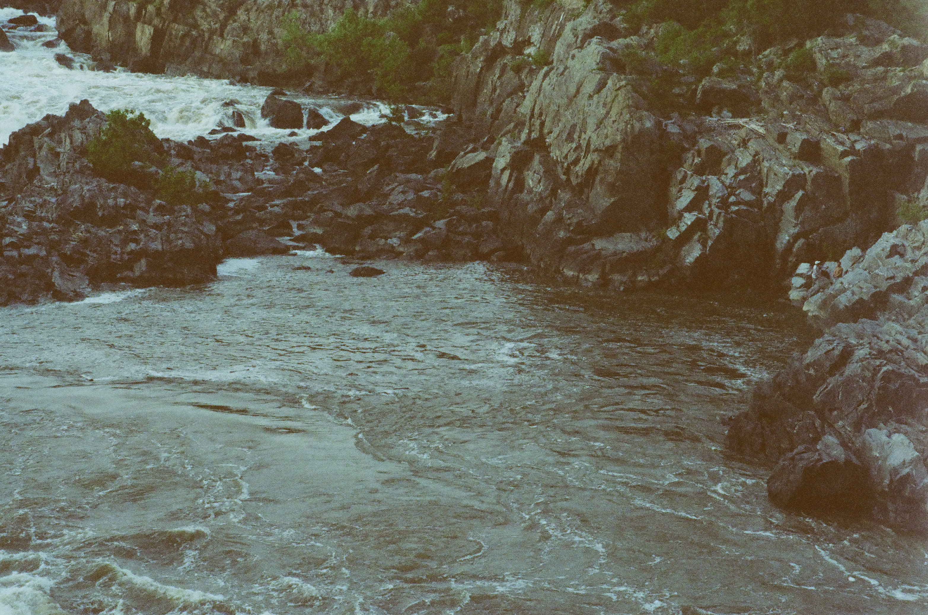 Whitewater churns between dark angular rocks at the top of the frame, feeding a calmer green pool where two people sit on the far bank.