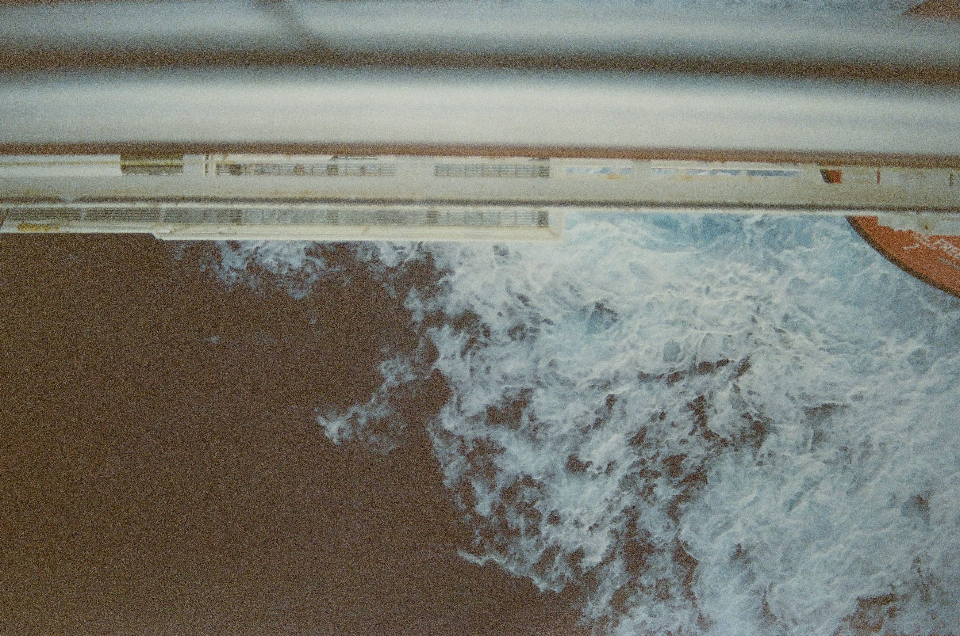 Blurred railing bars cross the frame above a long white ship structure, with churned white foam spreading over dark water below.