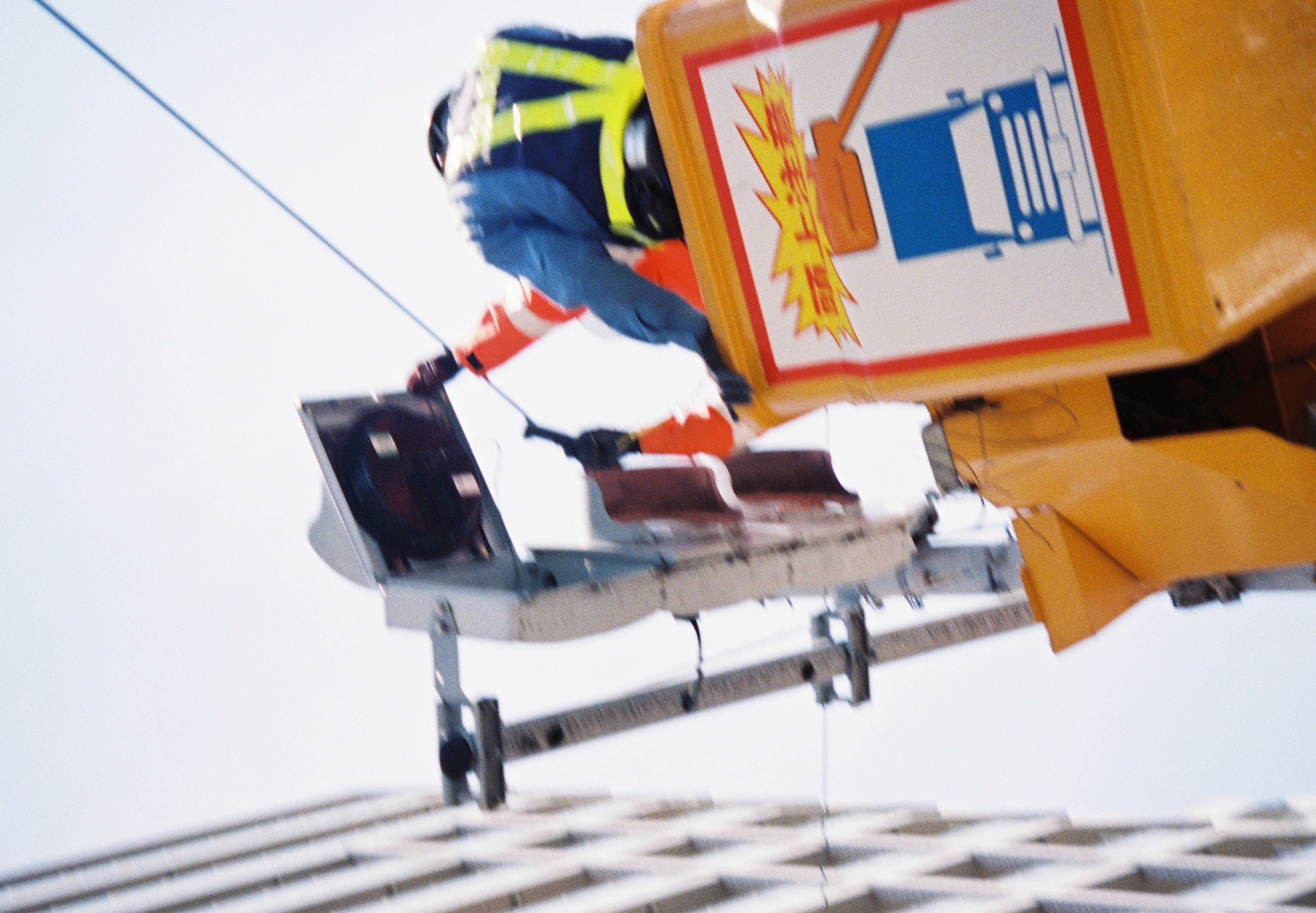 A worker in a hi-vis vest reaches from a yellow bucket lift toward a traffic signal head against a pale sky.