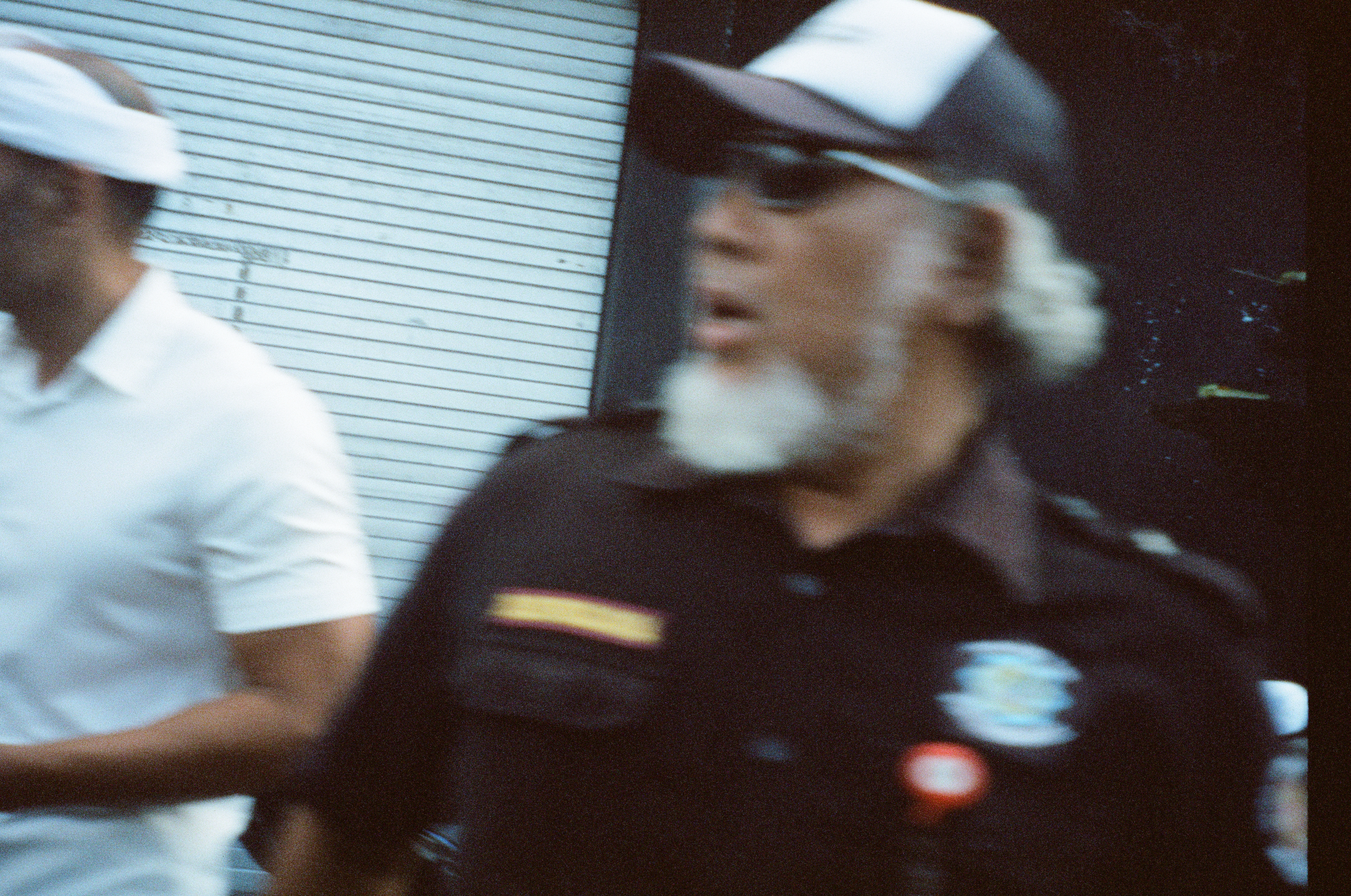 A white-bearded adult in sunglasses and a patched dark shirt fills the foreground beside a person in white against a metal shutter.