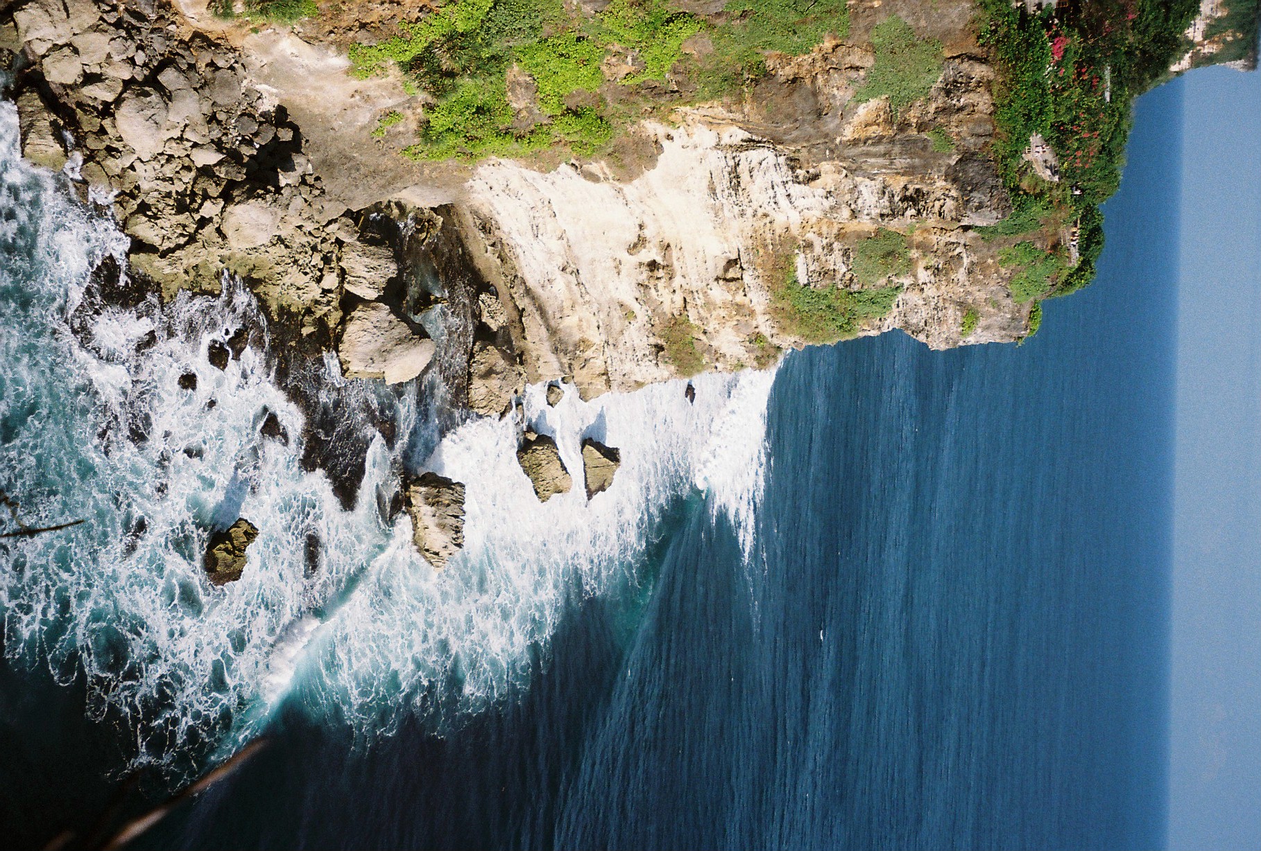 A vegetation-covered cliff drops toward scattered boulders as white surf curves along the deep blue water below.