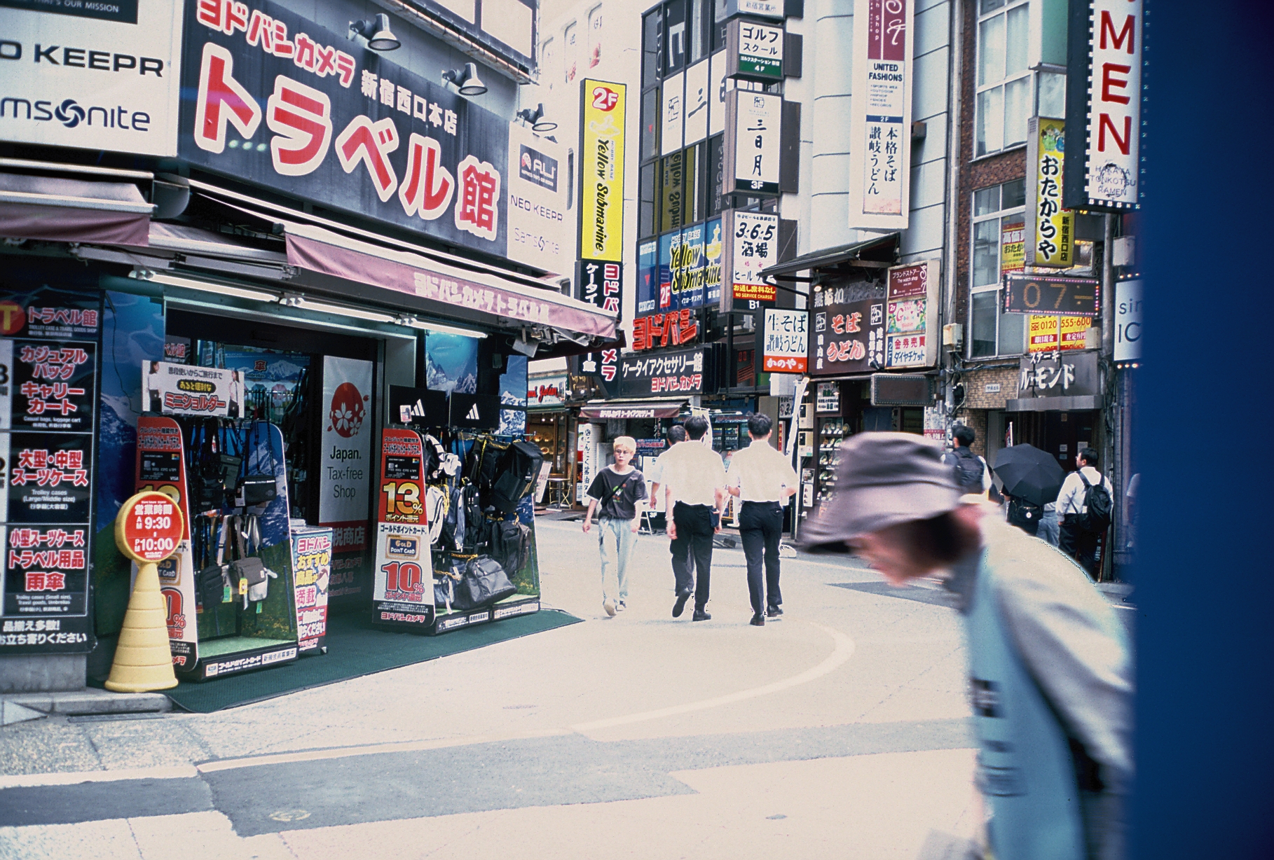 Pedestrians in white shirts walk down a paved street lined with dense vertical storefront signage and luggage displays in daylight.