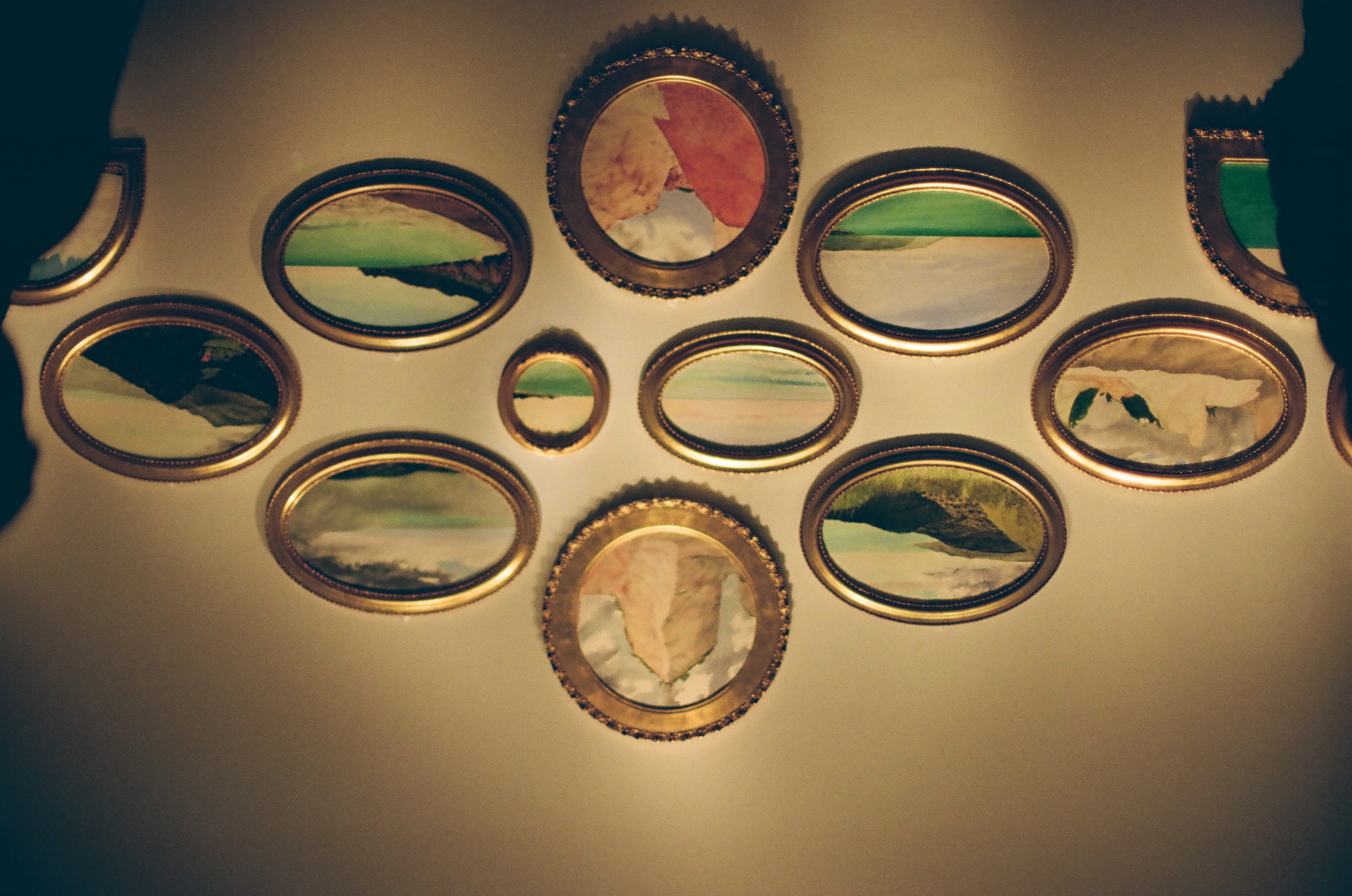 Twelve small oval and round landscape paintings in gold frames hang in a diamond cluster on a pale wall, with two dark figures at the edges.