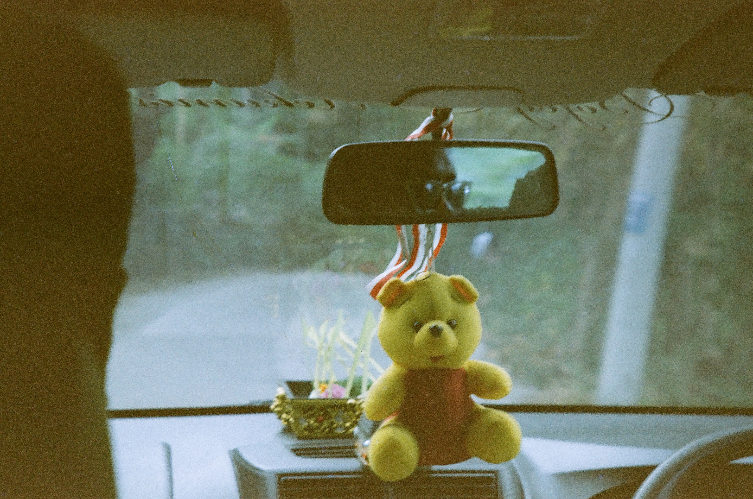 A yellow plush bear is positioned beneath a rearview mirror above a dashboard, with multicolored ribbons and a pale windshield view.