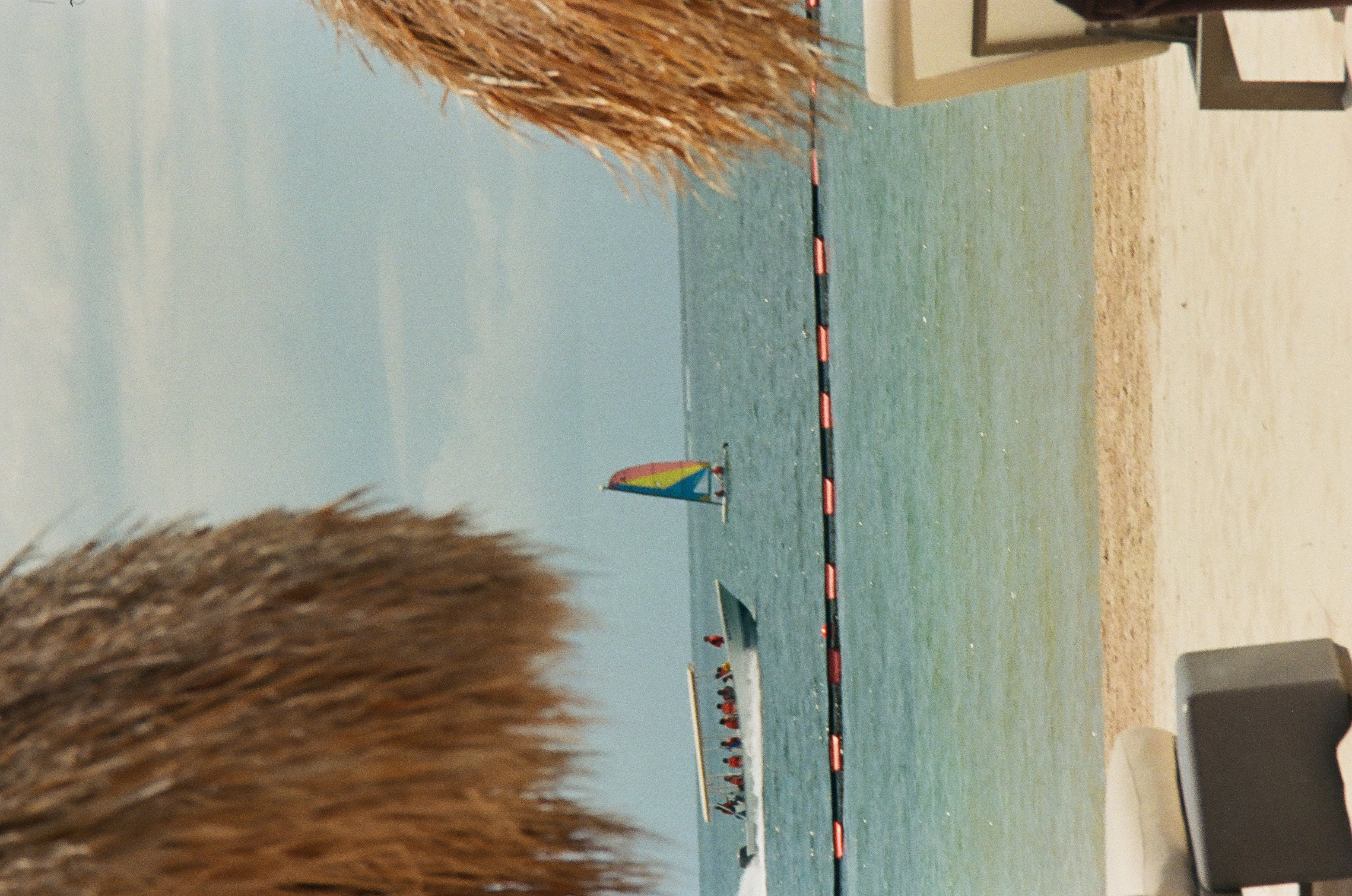A covered boat carrying several people and a smaller vessel with a multicolored sail appear beyond a segmented line of floats.