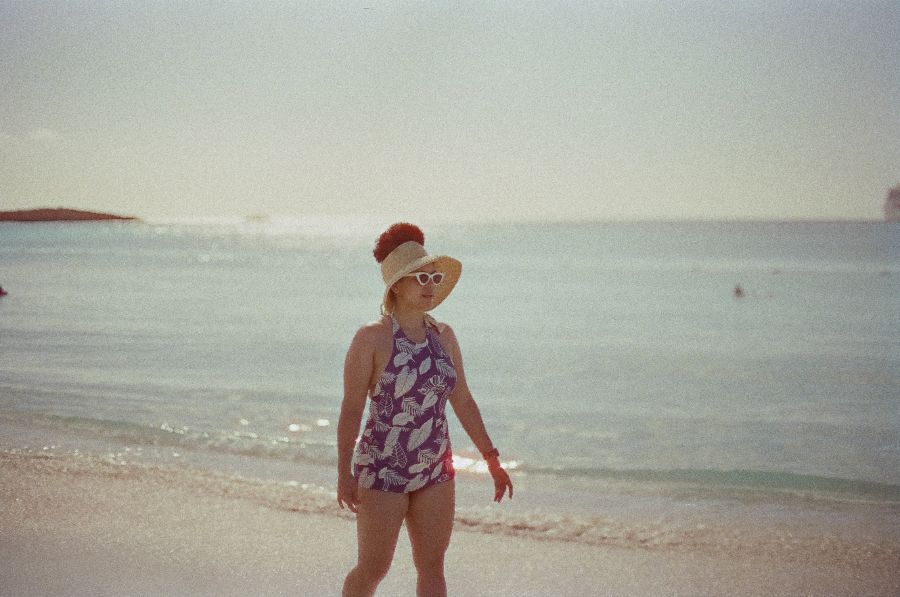 An adult wearing a broad hat, white sunglasses, and patterned swimsuit stands on wet sand facing right beside calm water.