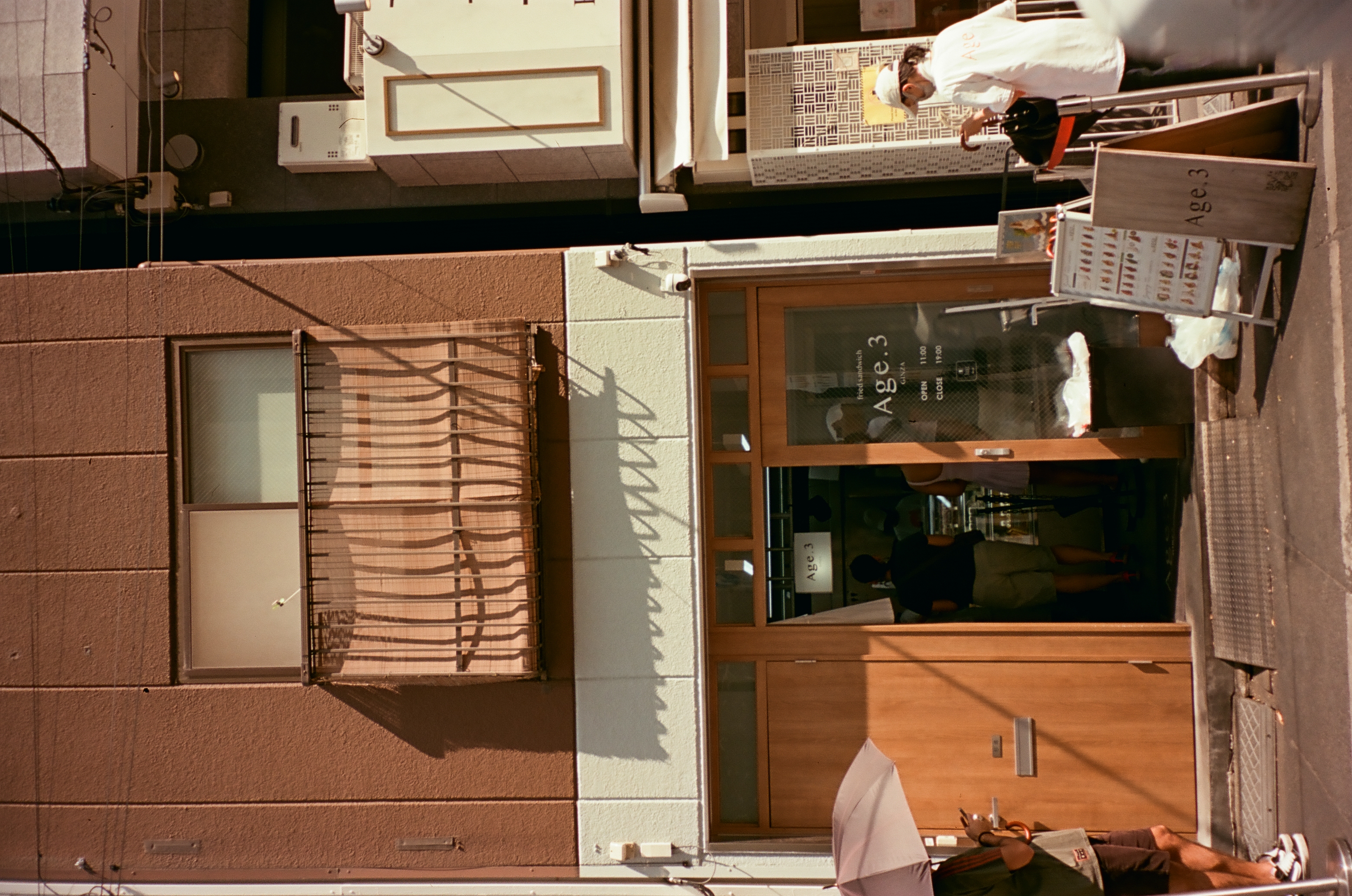 A narrow brown-tiled building with a slatted blind over its balcony, wooden doors below, a sandwich board reading Age.3, and five people nearby.
