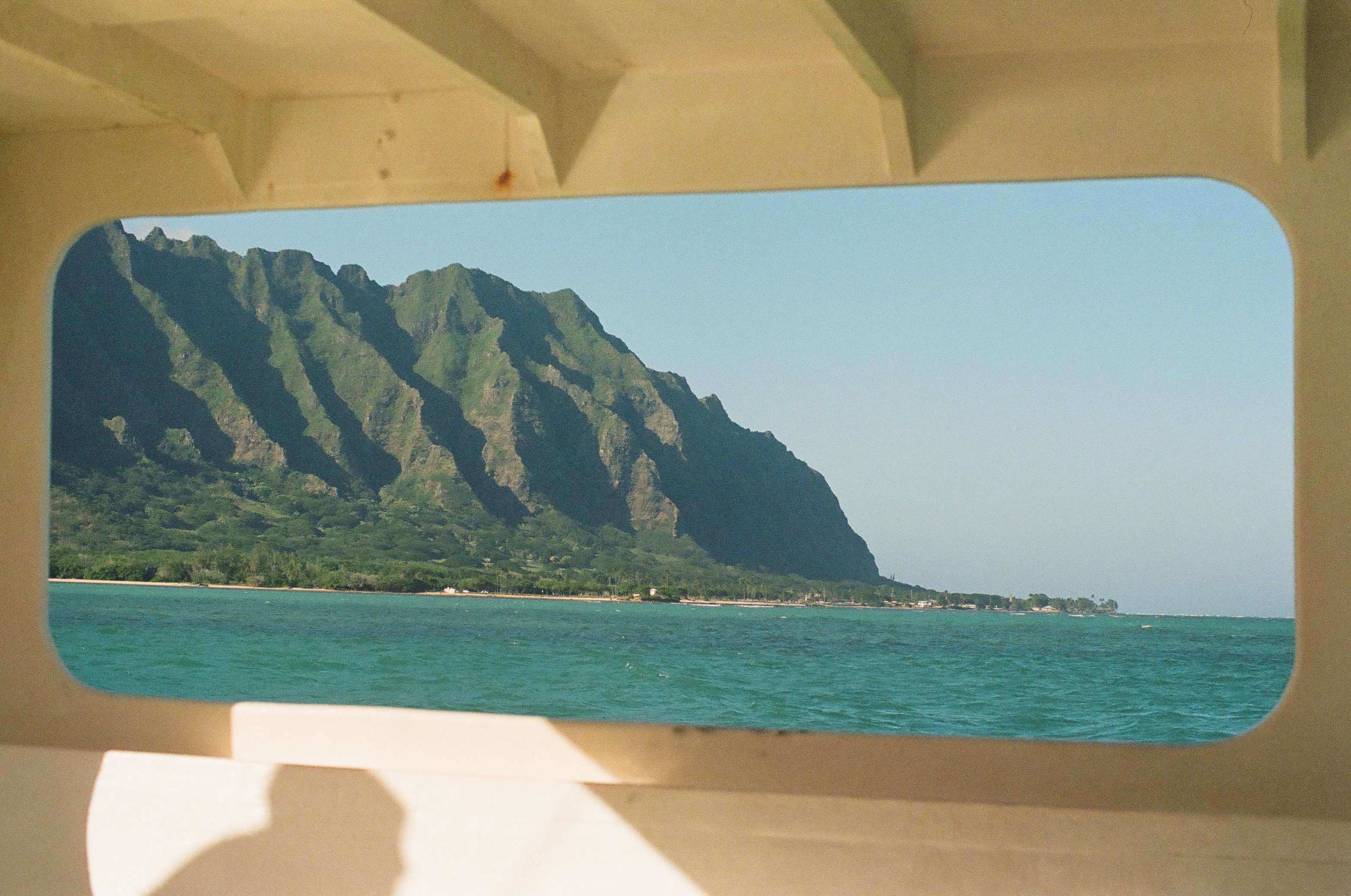 A rounded rectangular window in a boat's tan interior frames the same green mountain ridge and pale shoreline beyond calm blue-green water.