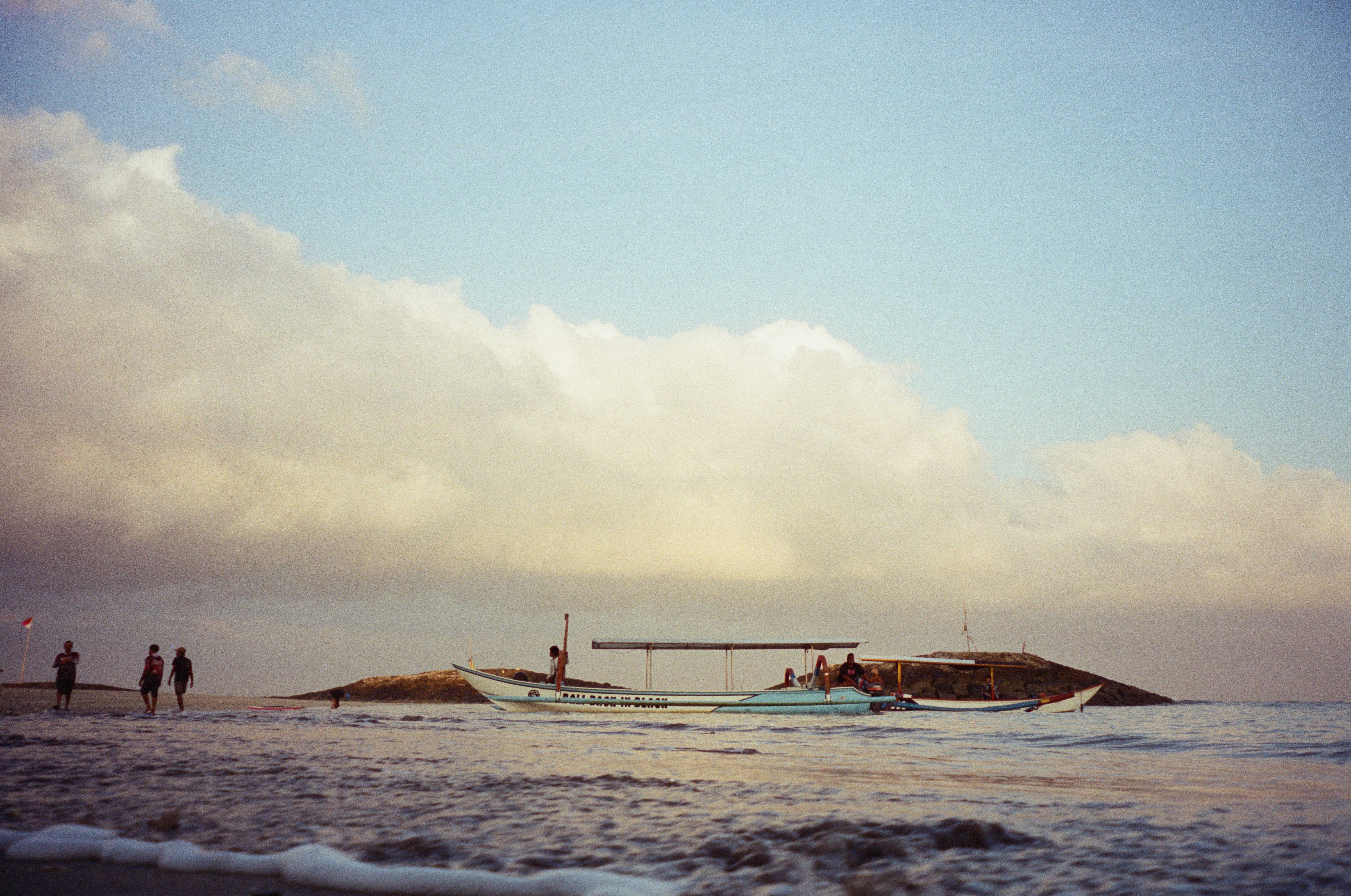 Two long canopied boats float near a rocky point while several people stand or wade beneath a broad bank of clouds.