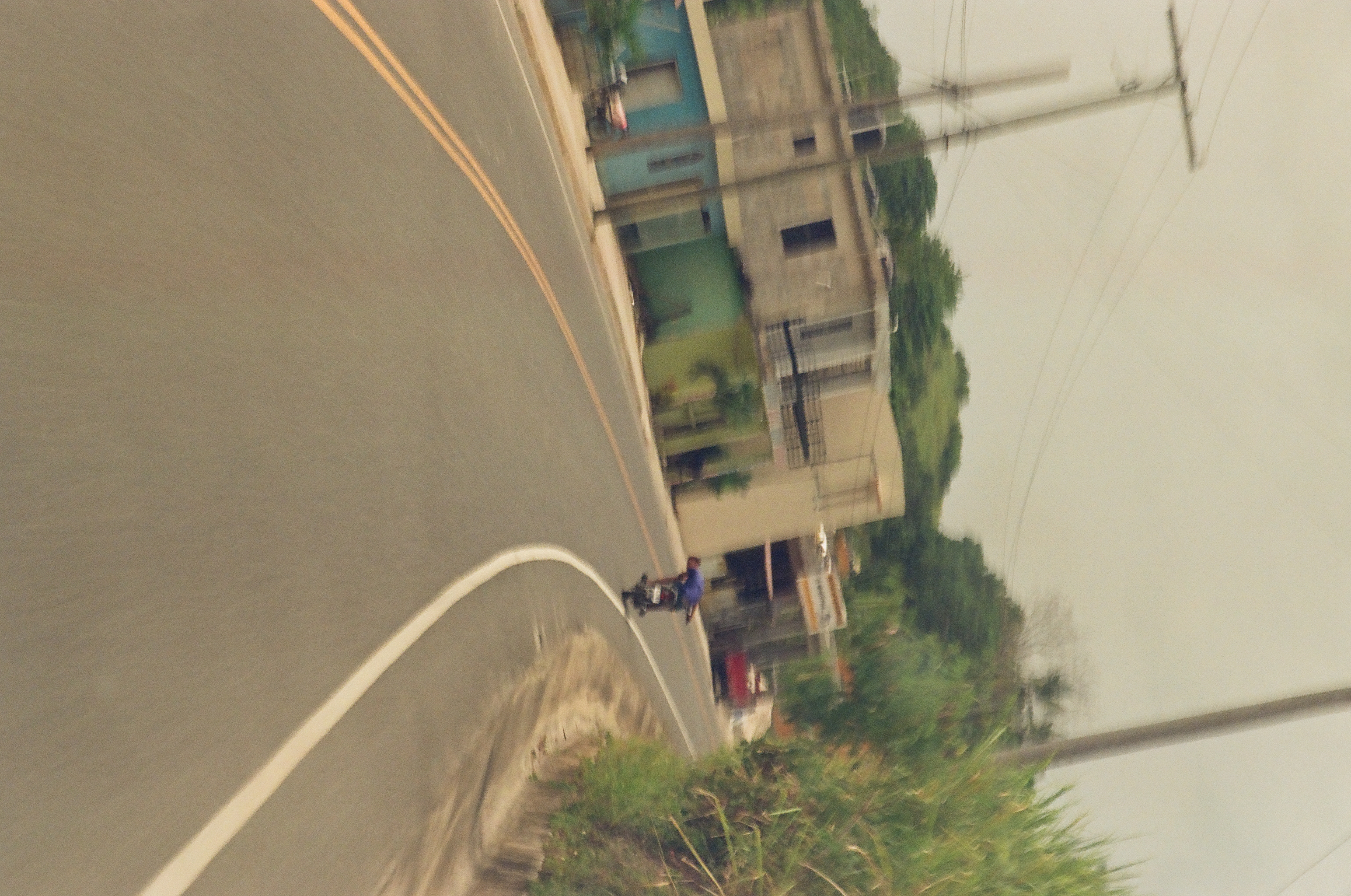 A motorcycle rider in a blue shirt follows a curving two-lane road past blue and green buildings, utility poles, and tall grass.