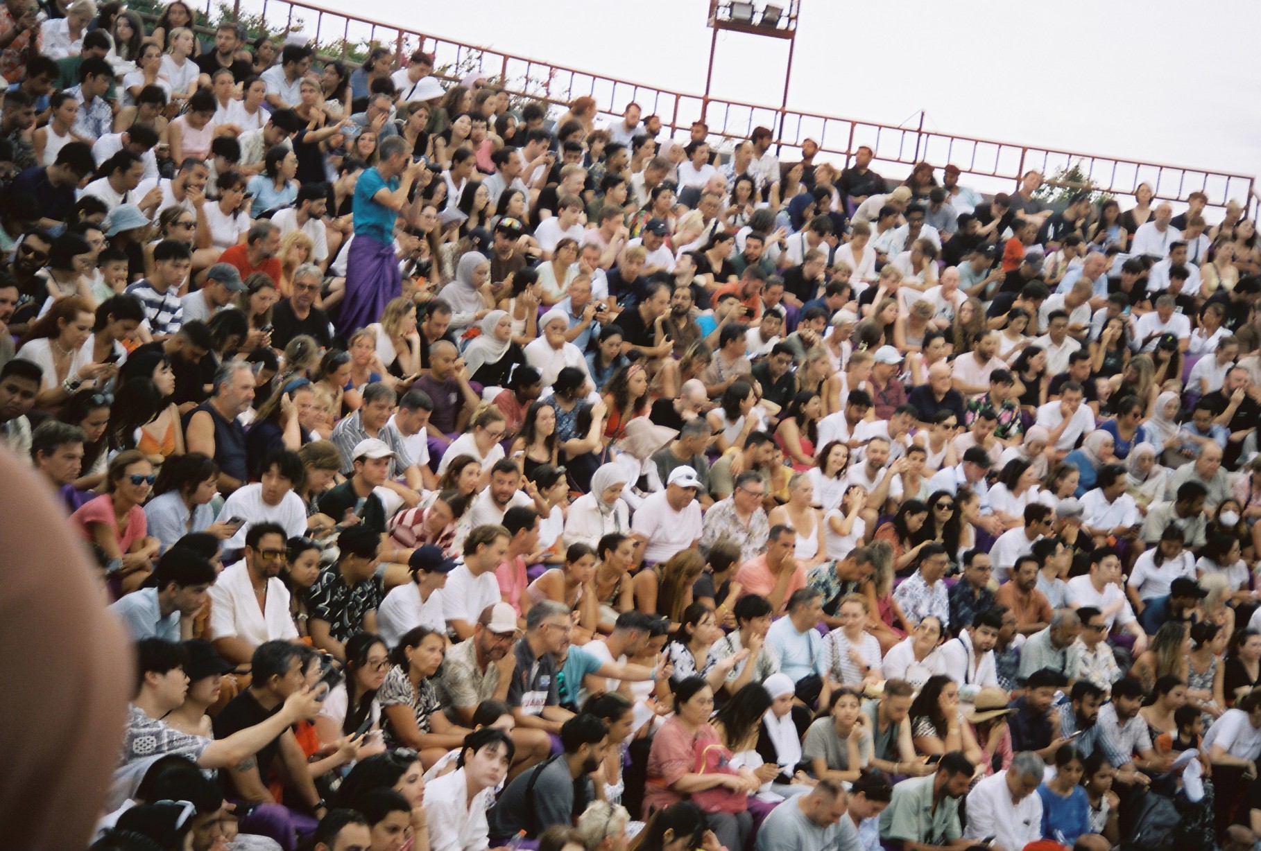 A tightly packed audience fills steep rows, with varied faces, clothing, and gestures visible across the bright crowd.