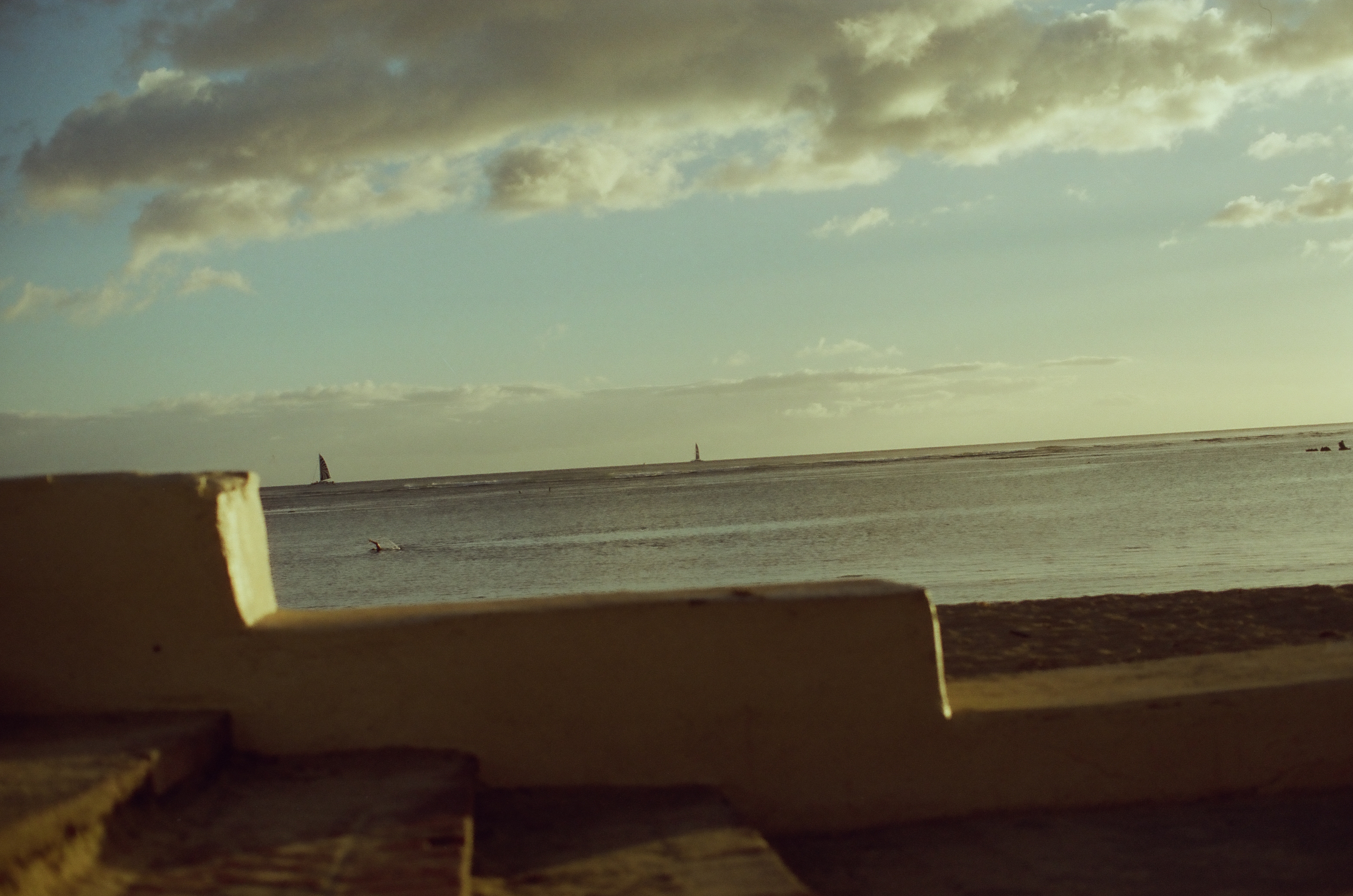 A cropped low wall and shadowed pavement frame pale water, warm sky, and several tiny sails on the horizon.