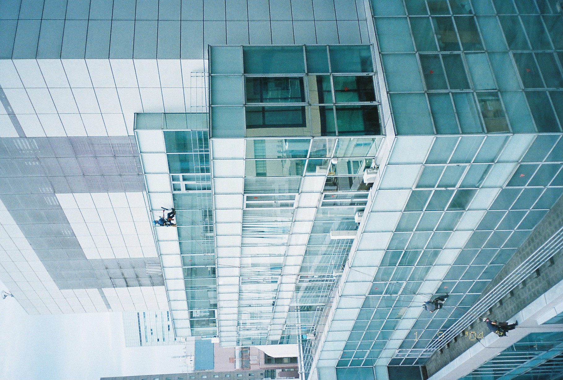 Three window washers on ropes work down the stepped glass-and-steel corner of an office building in cool blue-green light.