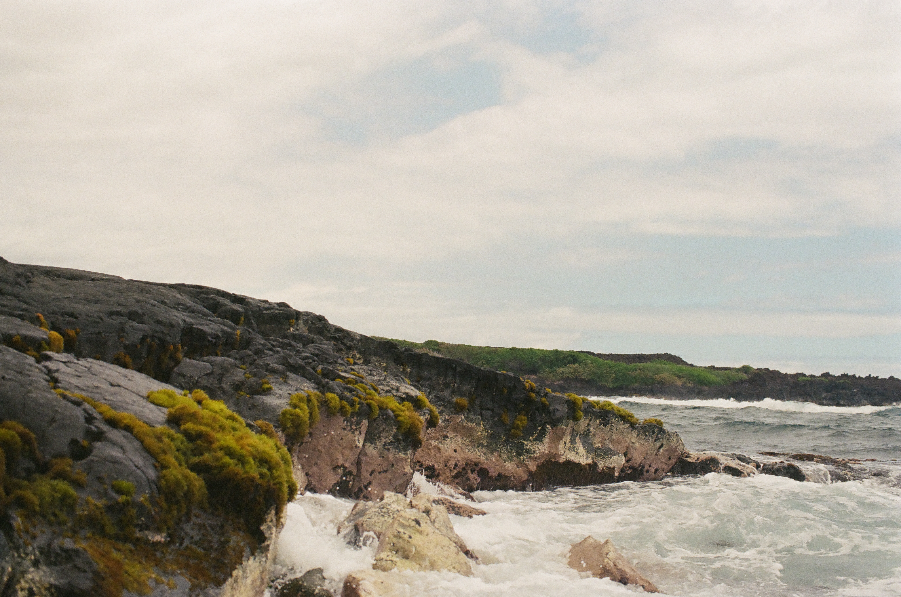 A mossy black rock ledge curves along the coast as foam recedes from its base, with distant waves breaking near a green headland.