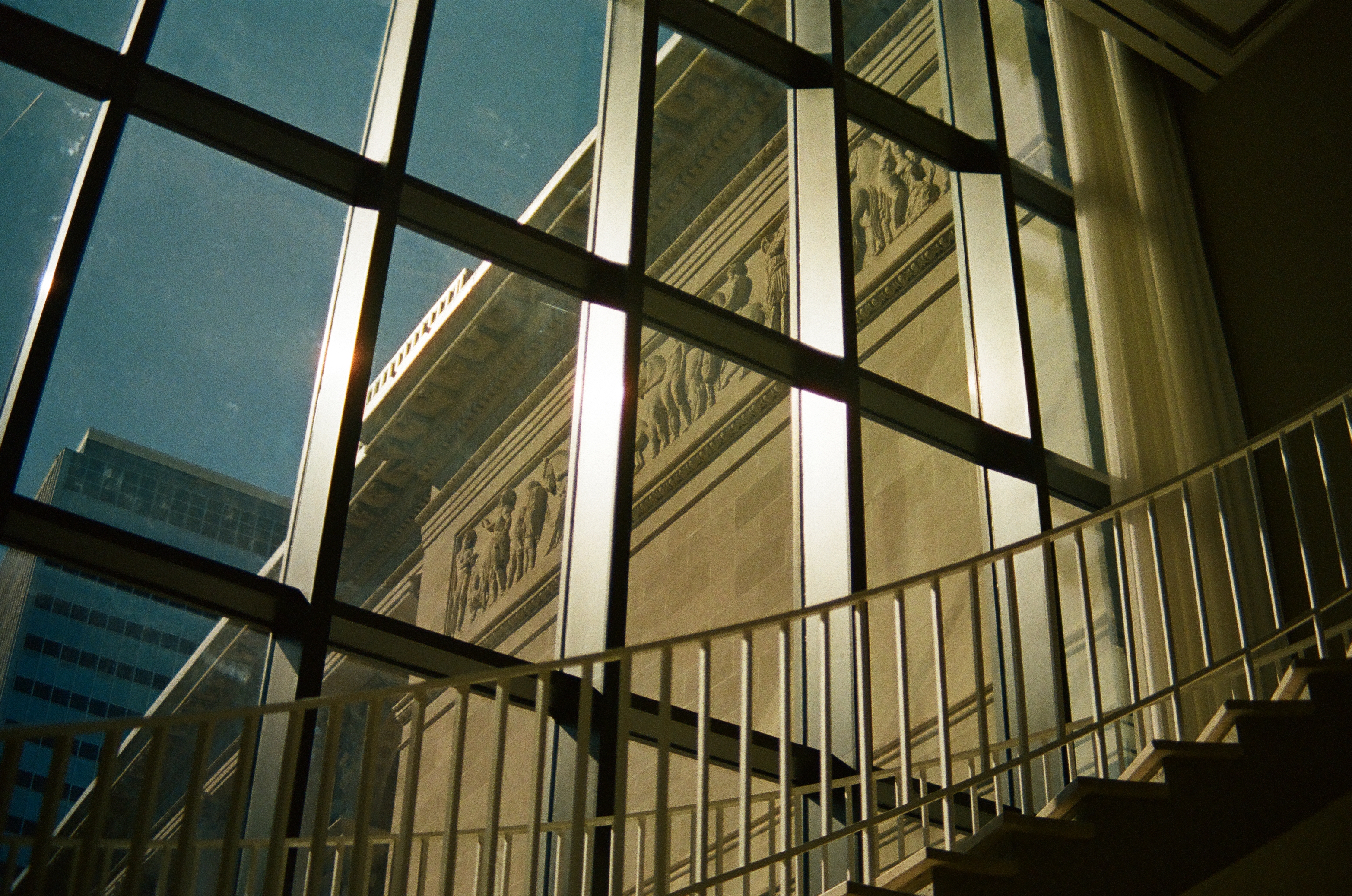 Window mullions and a stair railing frame a sunlit stone facade reflected across dark glass.