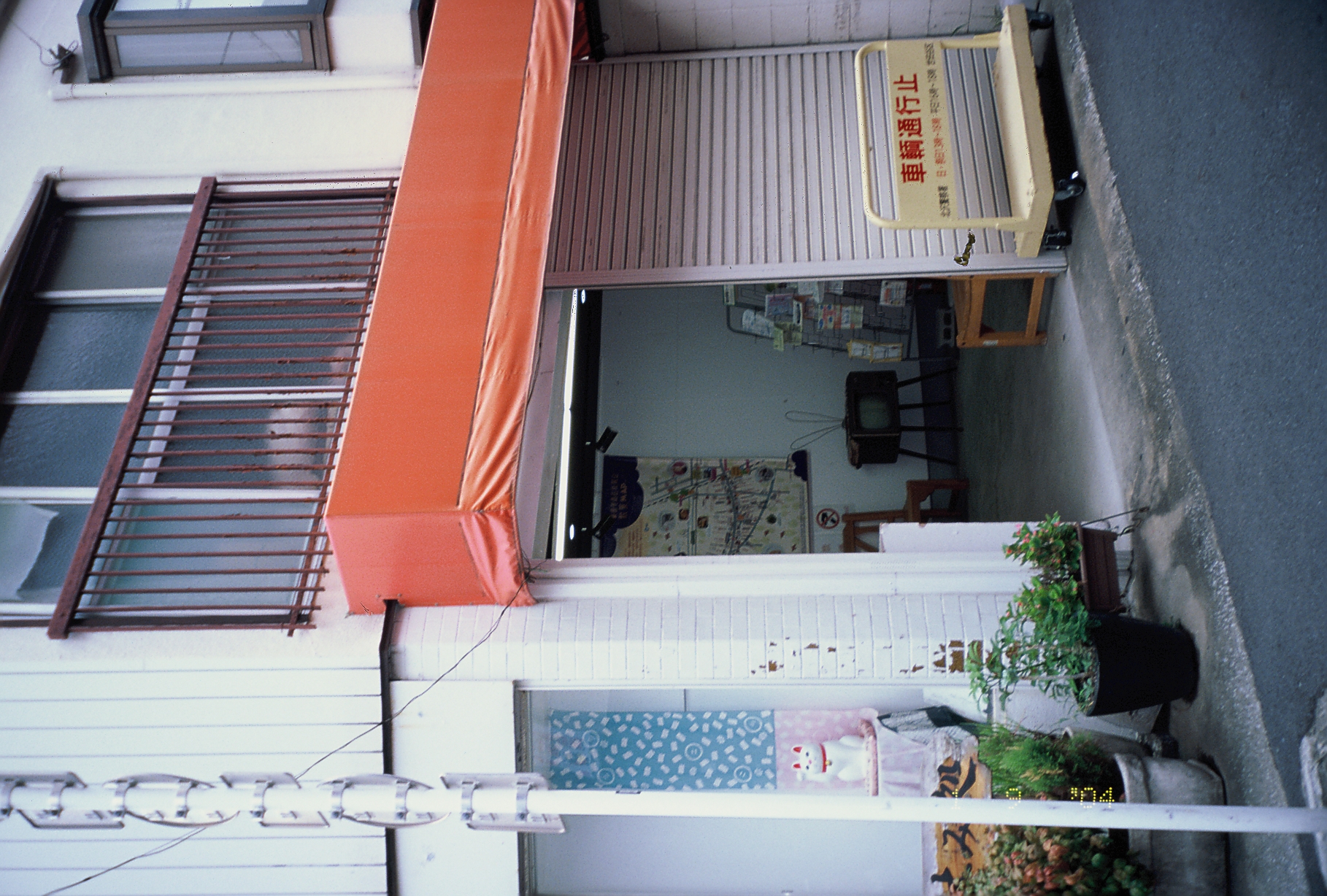 A bright orange awning shades an open storefront doorway, with potted plants on the step and a wheeled yellow barrier at the curb.