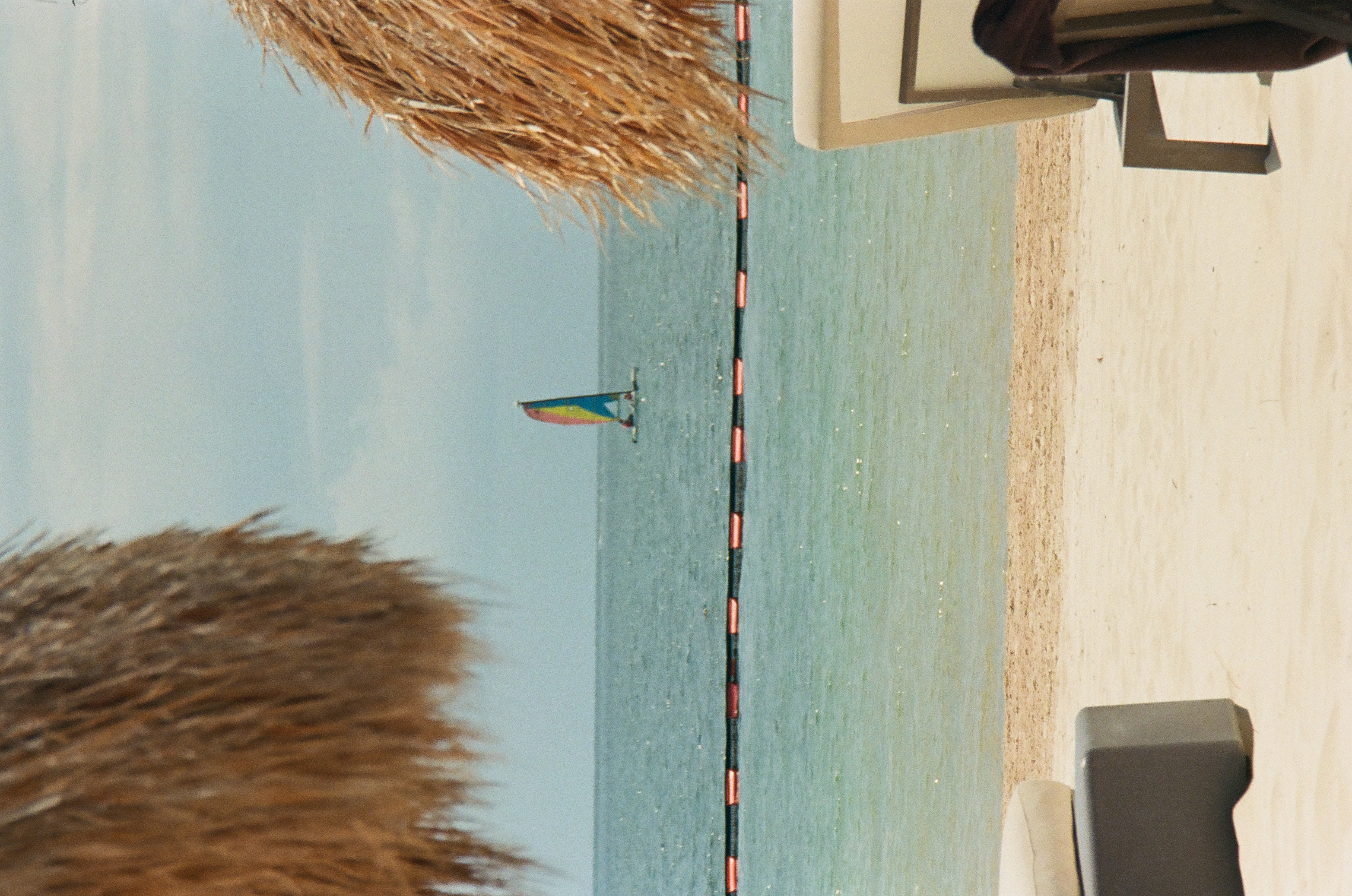 Thatched fringes frame pale sand, blue-green water, a segmented line of floats, and a distant vessel with a multicolored sail.