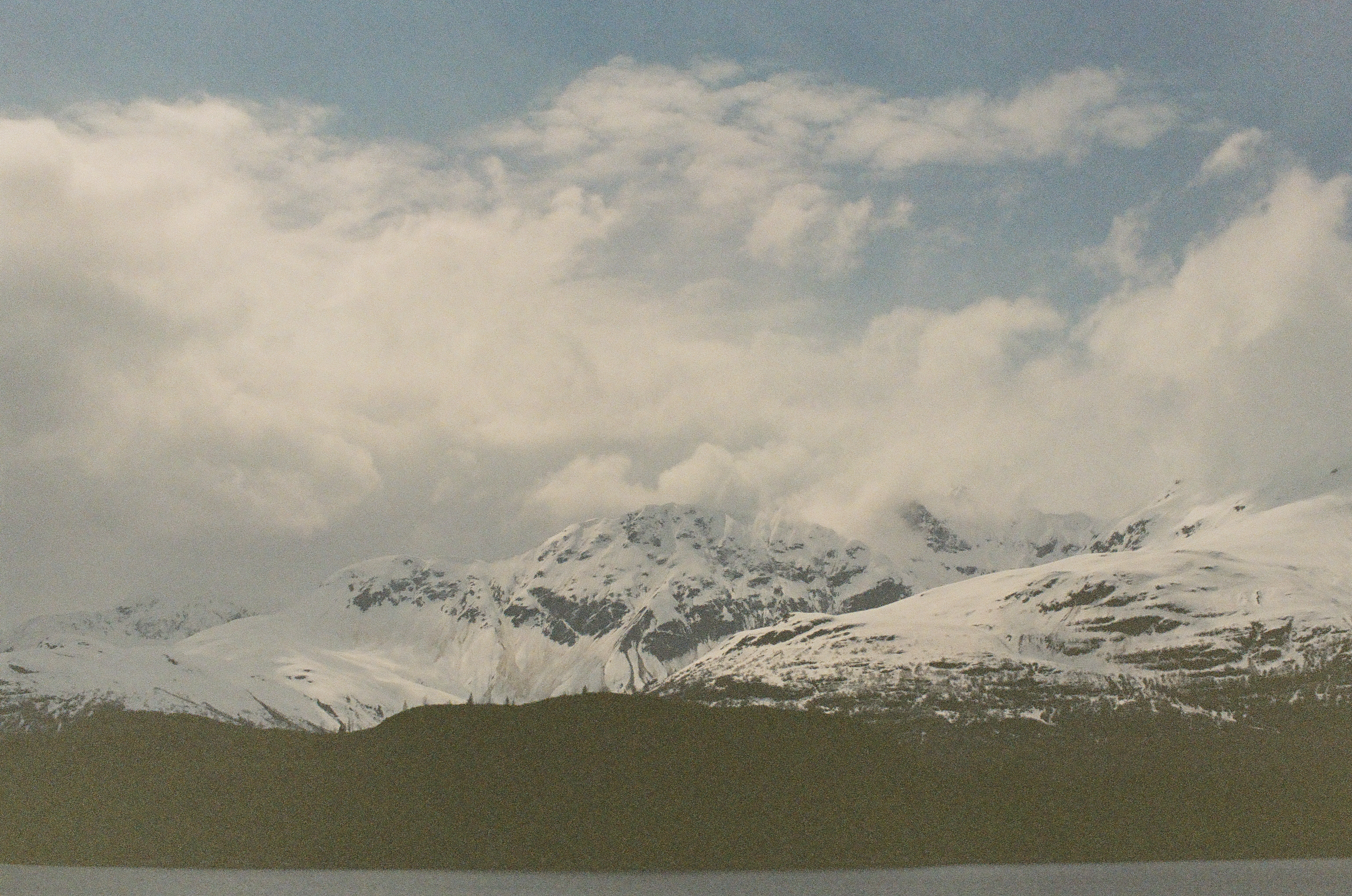 Jagged white ridges extend behind a dark shoreline beneath broken clouds and a pale blue sky.