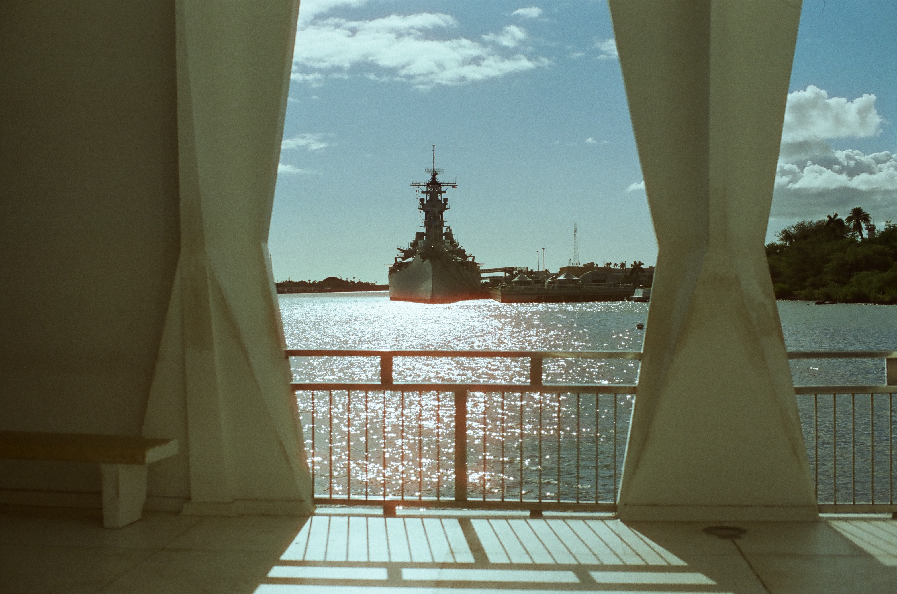 White structural openings frame a gray naval vessel across bright water beneath scattered clouds and blue sky.