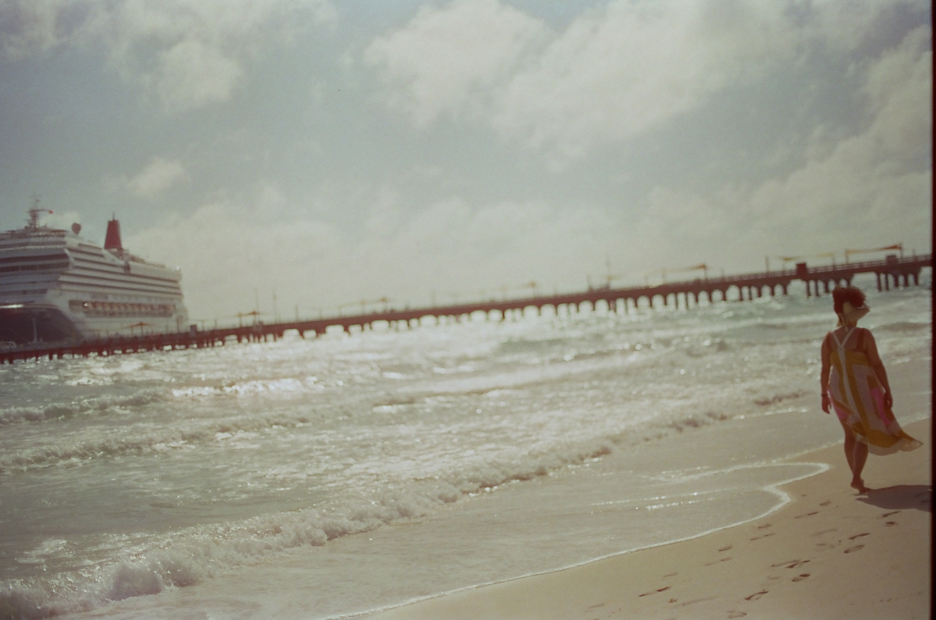 A person in a flowing patterned dress walks along wet sand past footprints while a long pier crosses the surf toward a large white ship.