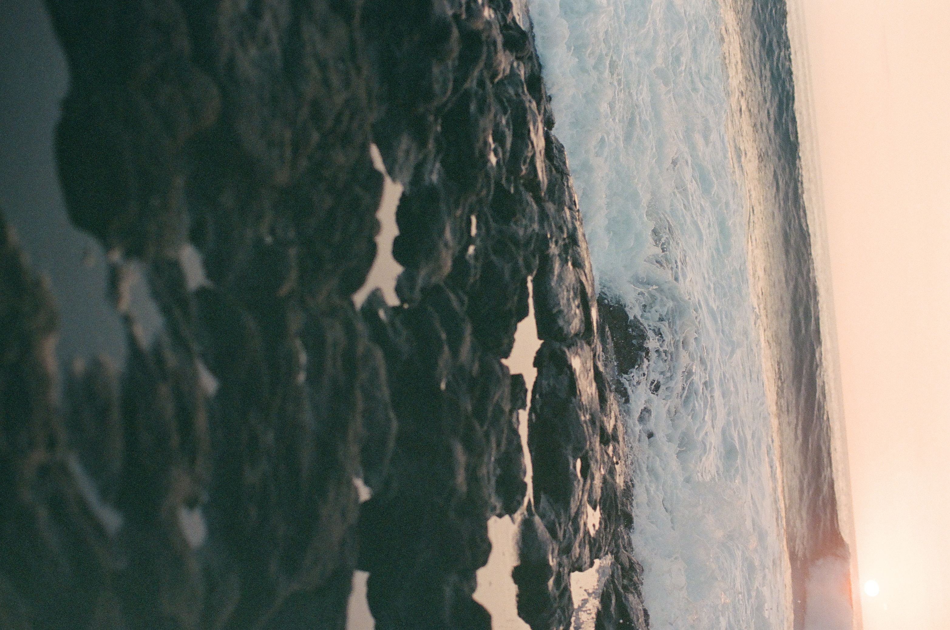 White surf pours through jagged dark rocks and shallow pools beneath a pale peach evening sky.