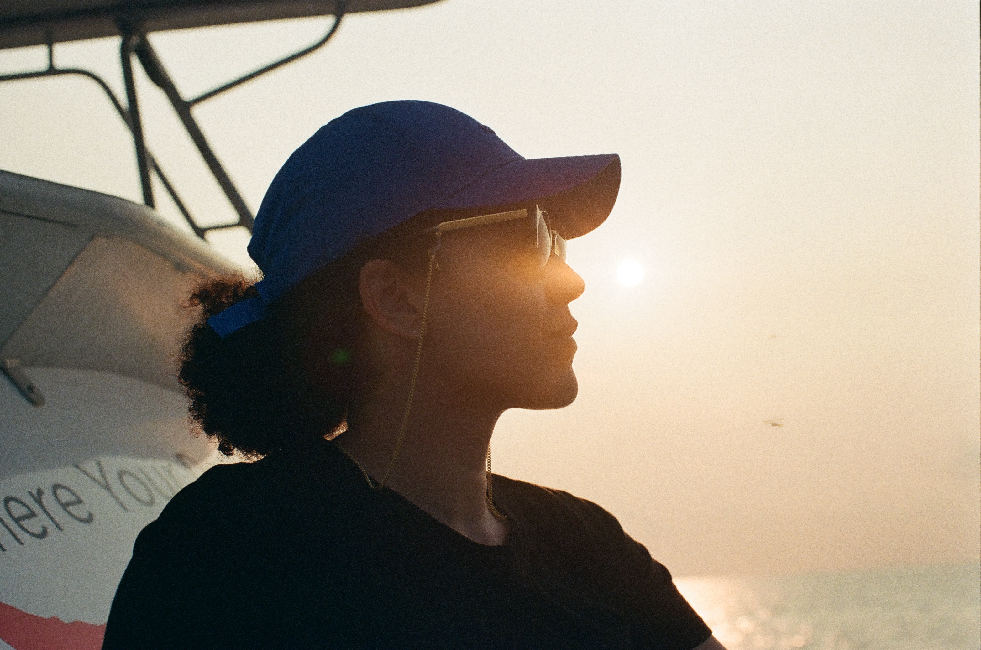 A person in a blue cap turns toward a low bright sun above shimmering water and a pale horizon.