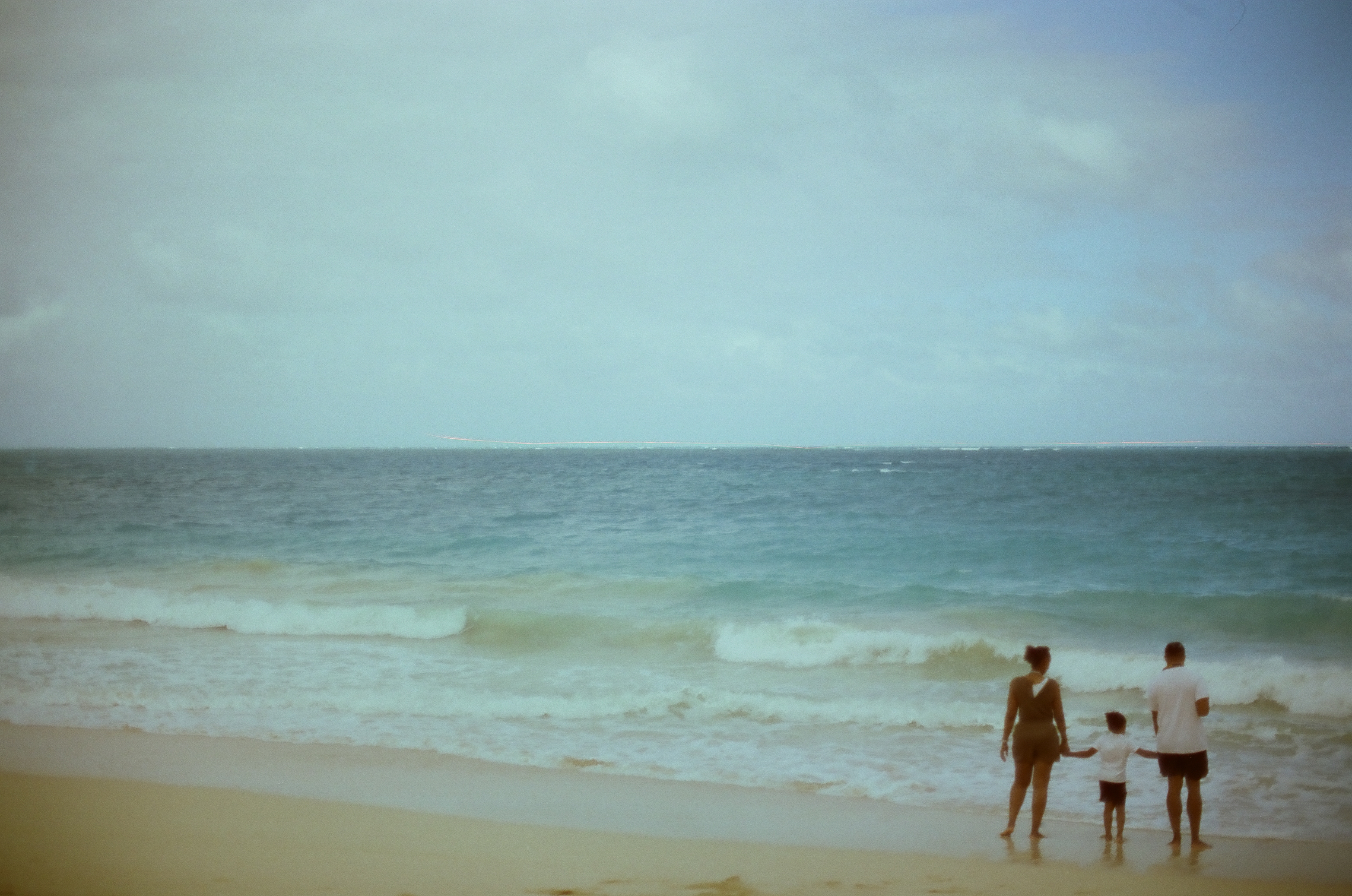 A shorter figure stands between two taller people holding hands at the edge of gentle breaking waves.