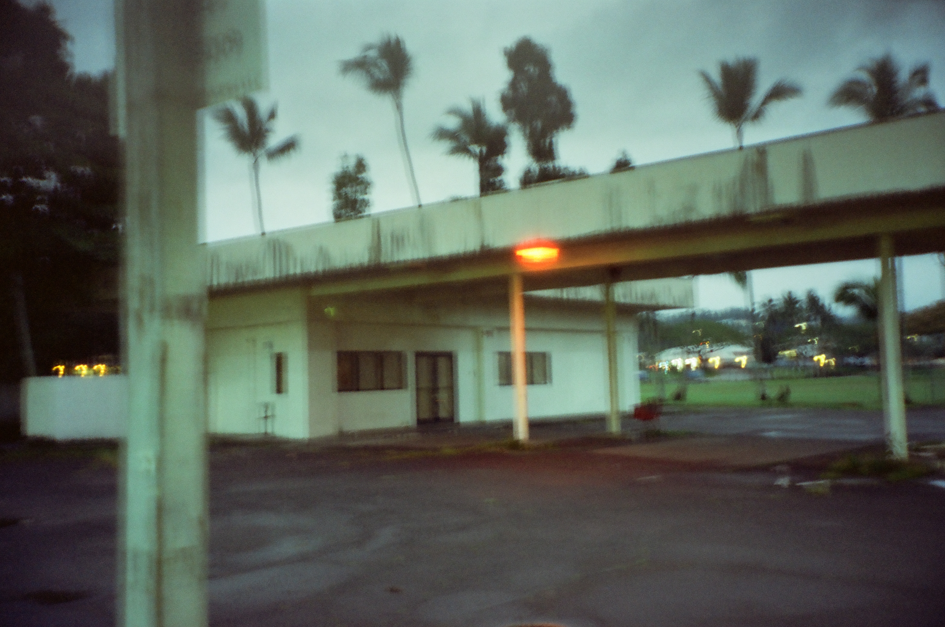 An empty low building and covered drive sit on wet pavement beneath blurred palms and evening light.
