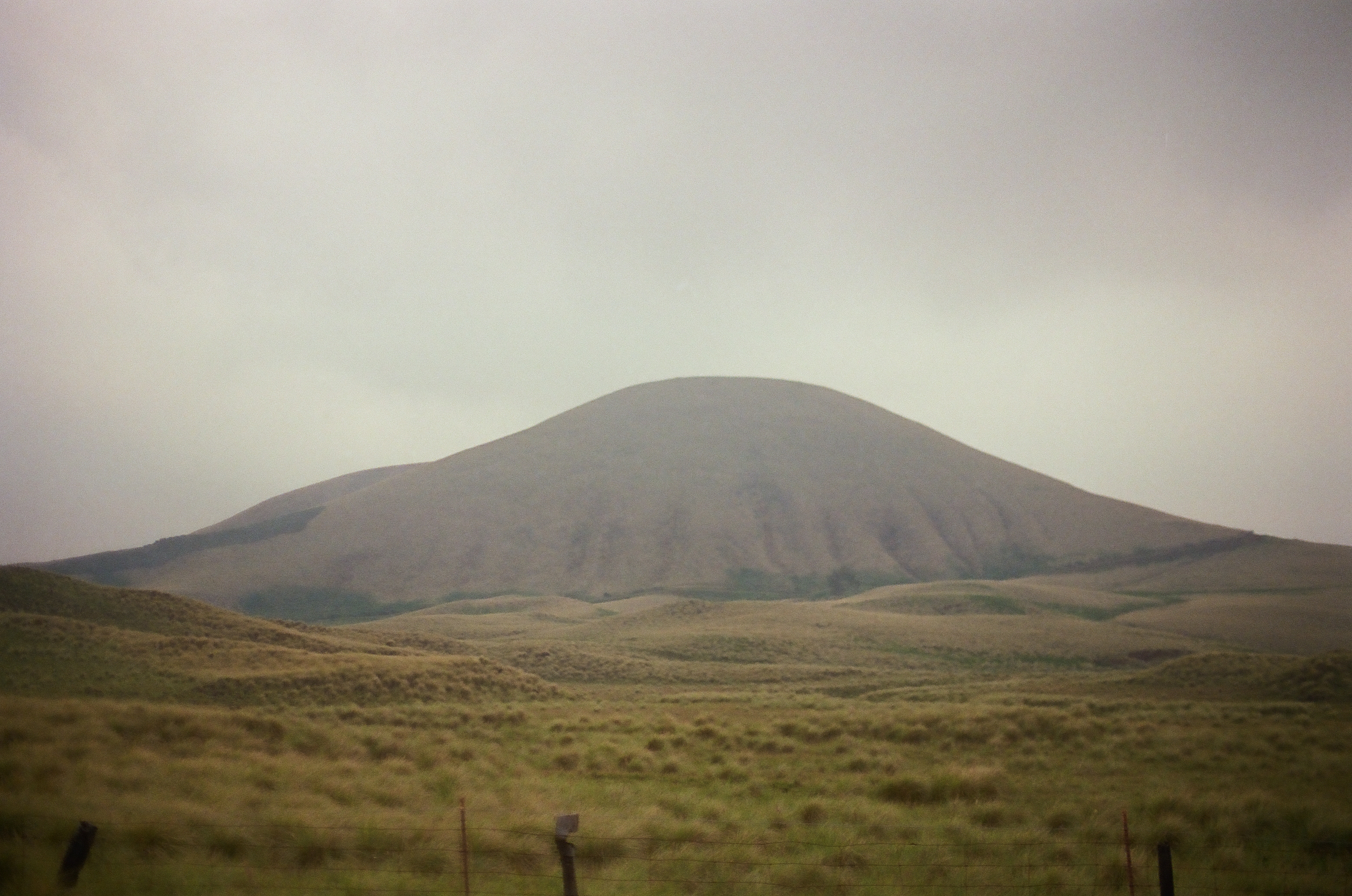 A broad rounded brown mountain rises from dry grass beneath a muted gray-blue sky.