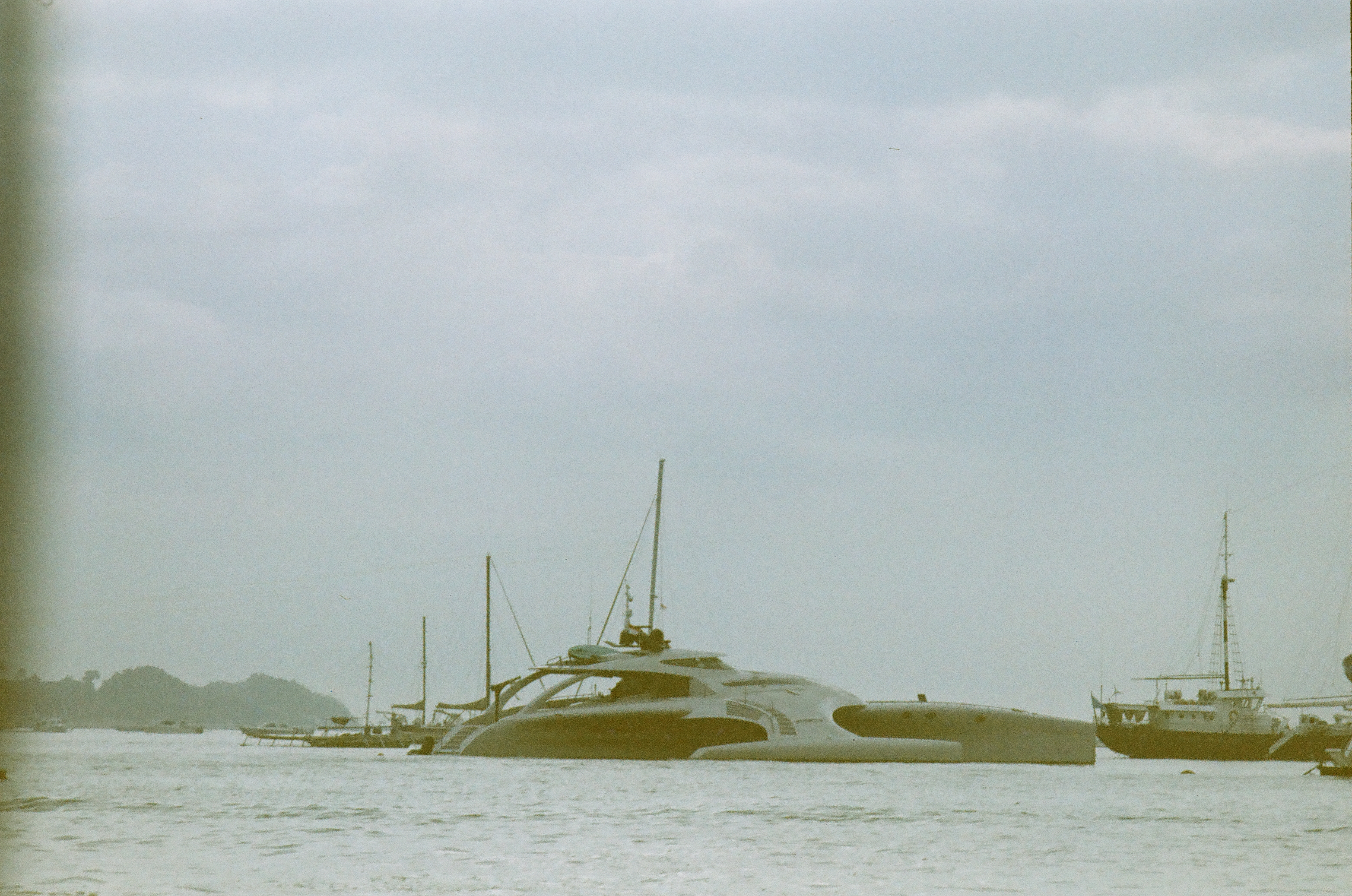A broad low yacht sits among several mast-bearing boats on muted water beneath a pale overcast sky.
