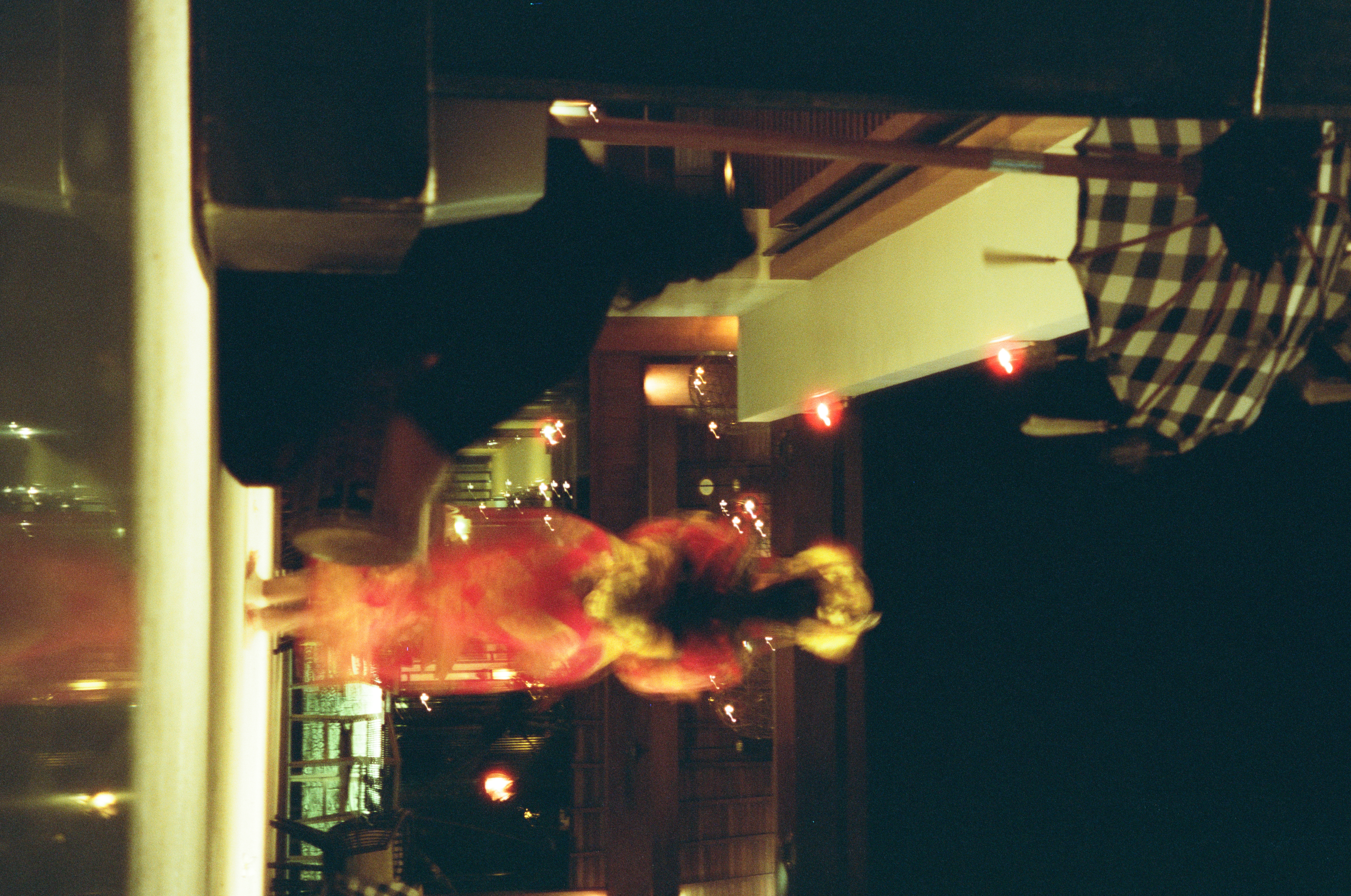 Beneath hanging checked fabric, a blurred red-and-gold figure with a large gold head adornment raises both arms in the distance beyond a dark seated silhouette.