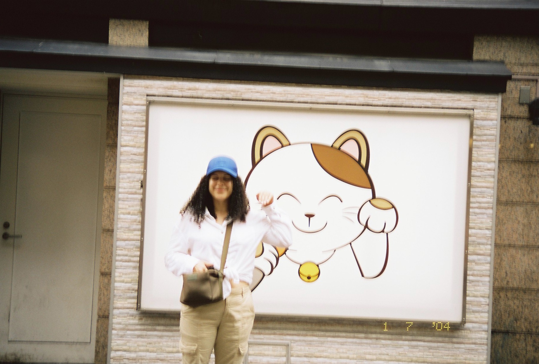 A person in a blue cap and white shirt smiles beside a large illustrated white and orange cat with a raised paw and a gold bell.