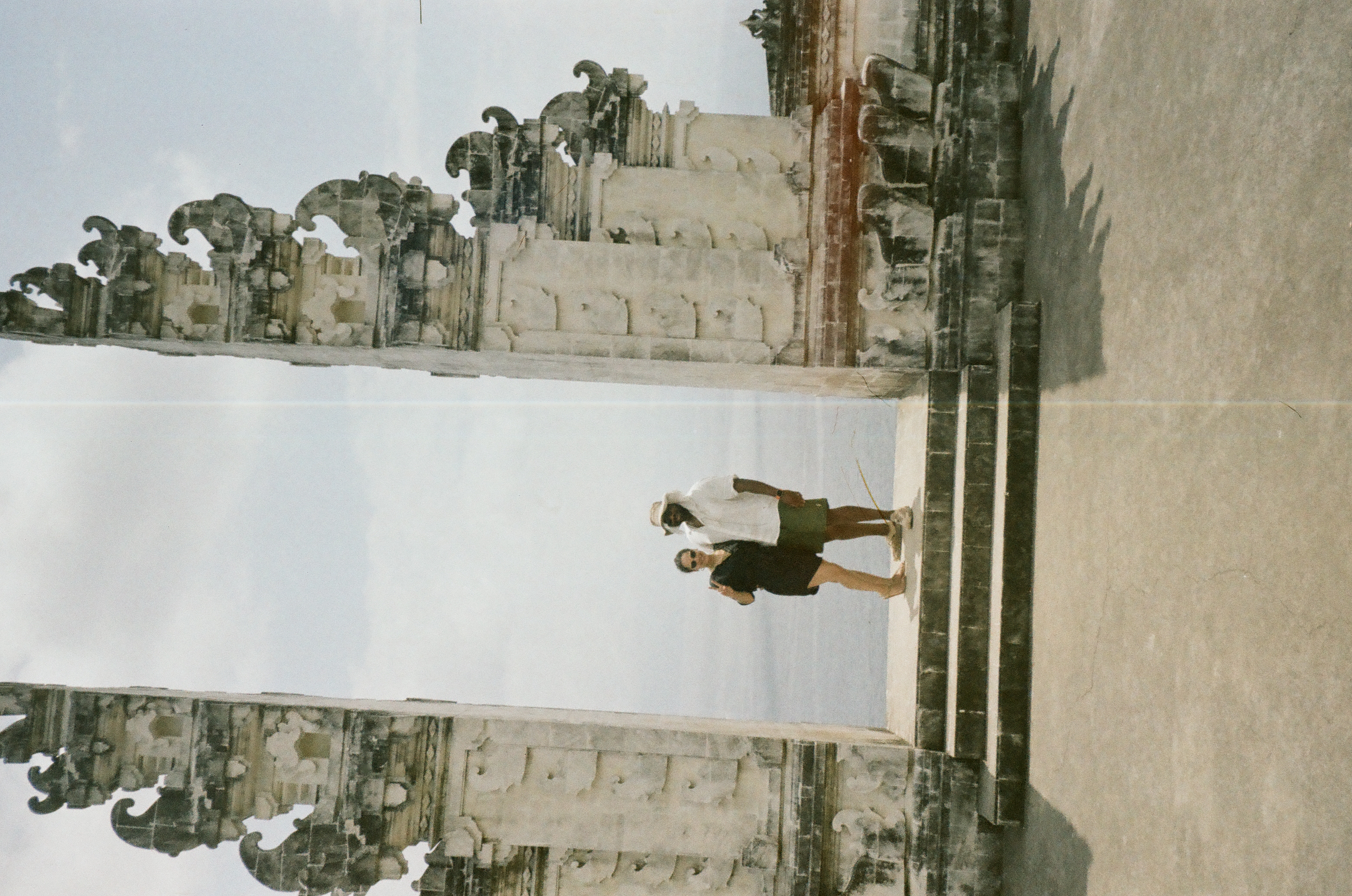 Two people stand on a raised threshold between weathered carved towers, with a pale vertical line running near the center.