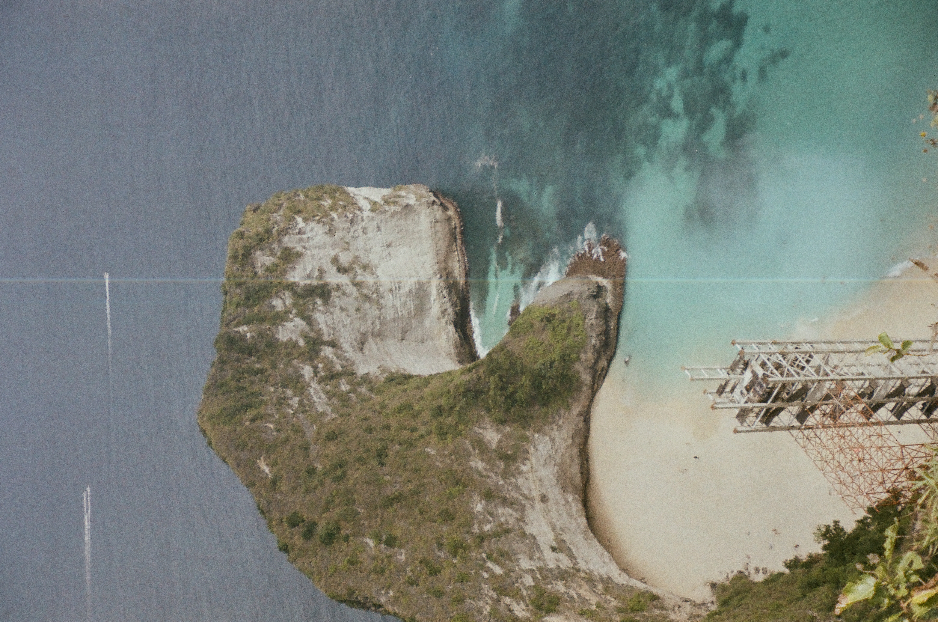A green headland curves around pale sand and turquoise shallows viewed from a high cliff.