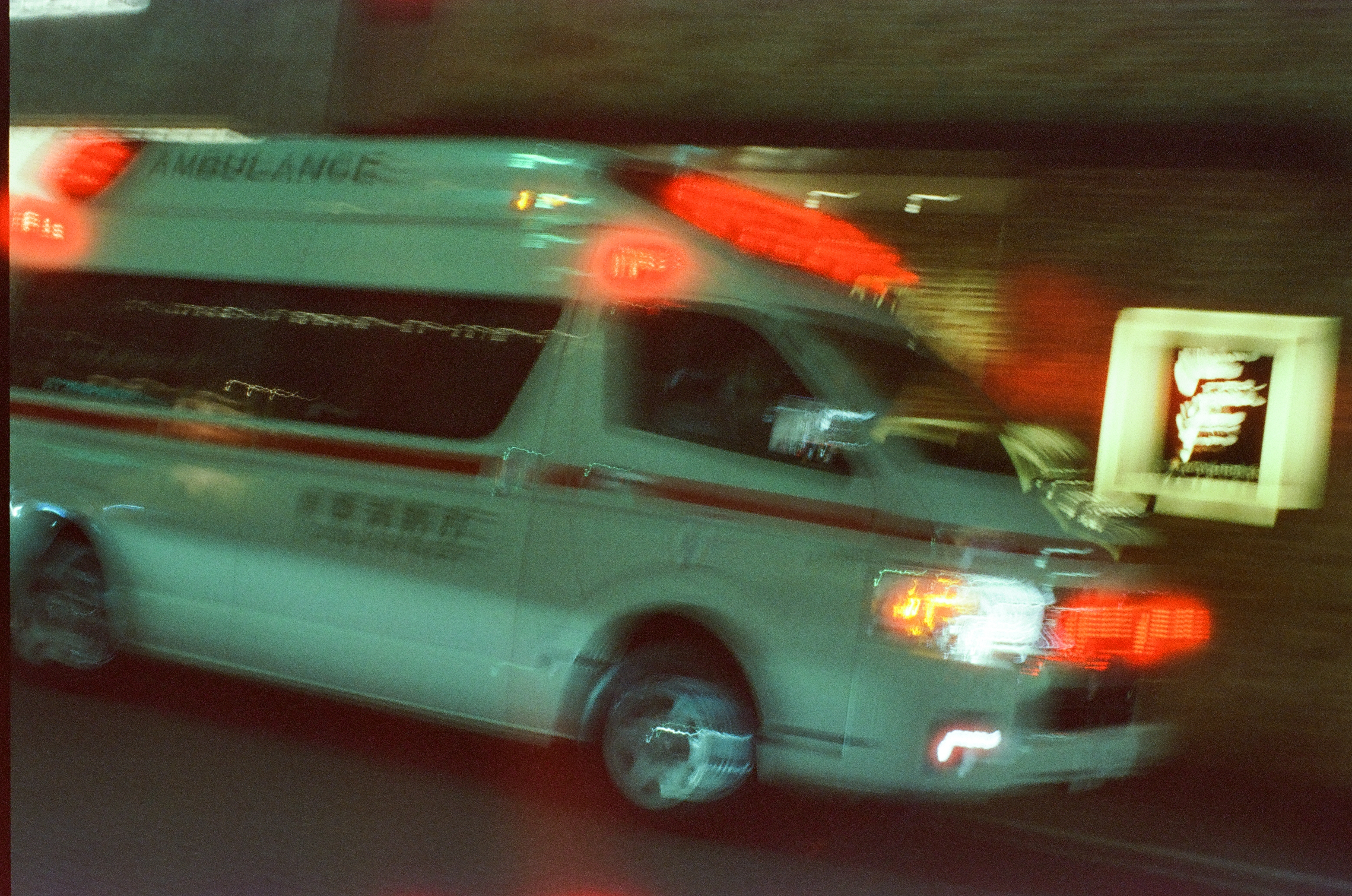 A motion-blurred white van marked AMBULANCE with red roof lights and a red side stripe passes a lit square sign against a dark background.