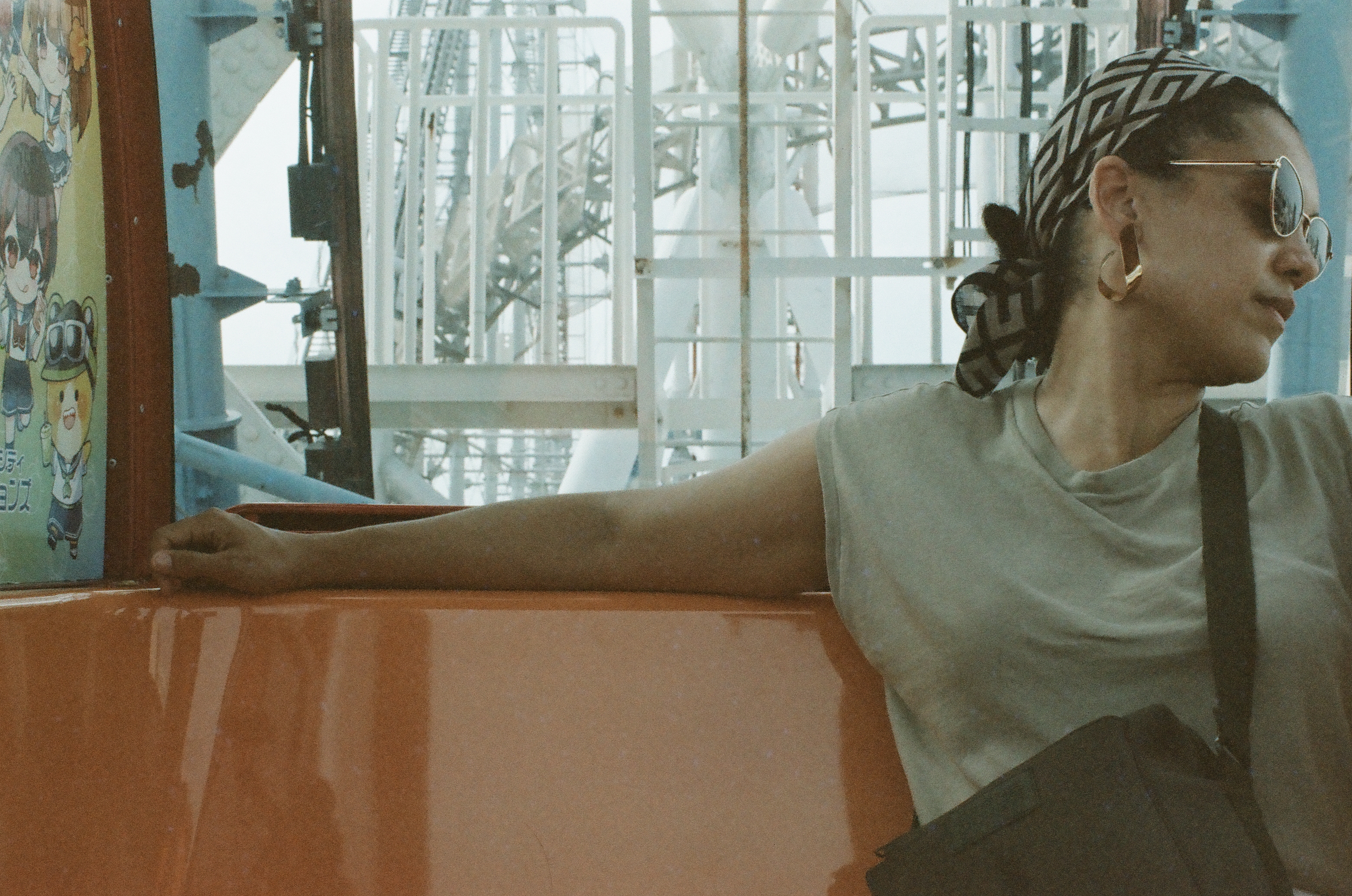 A woman in a patterned headscarf and round sunglasses rests her arm along an orange cabin sill, with pale steel Ferris wheel framework behind her.