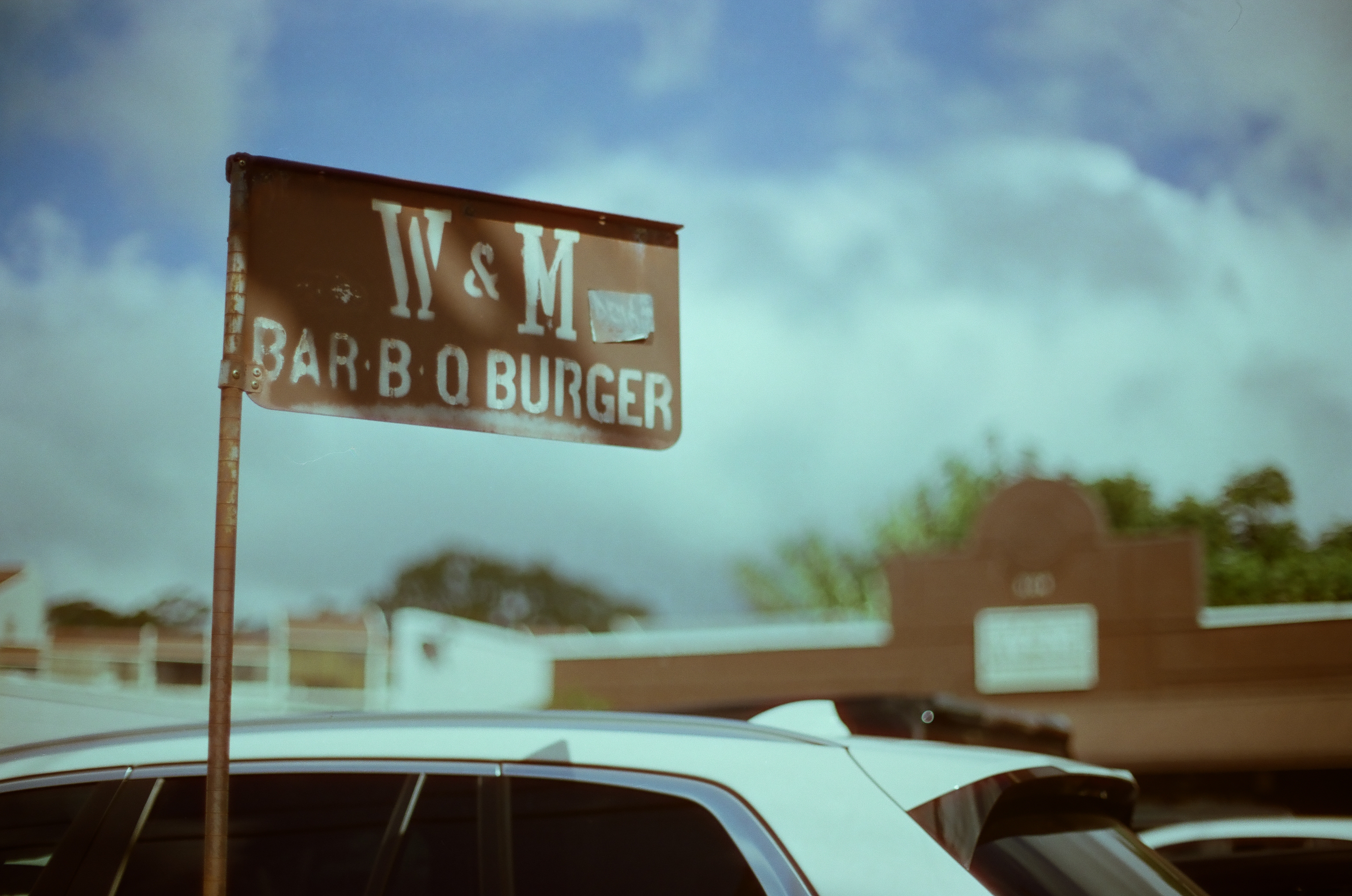 A weathered W and M Bar-B-Q Burger sign rises above parked cars beneath a clouded blue sky.