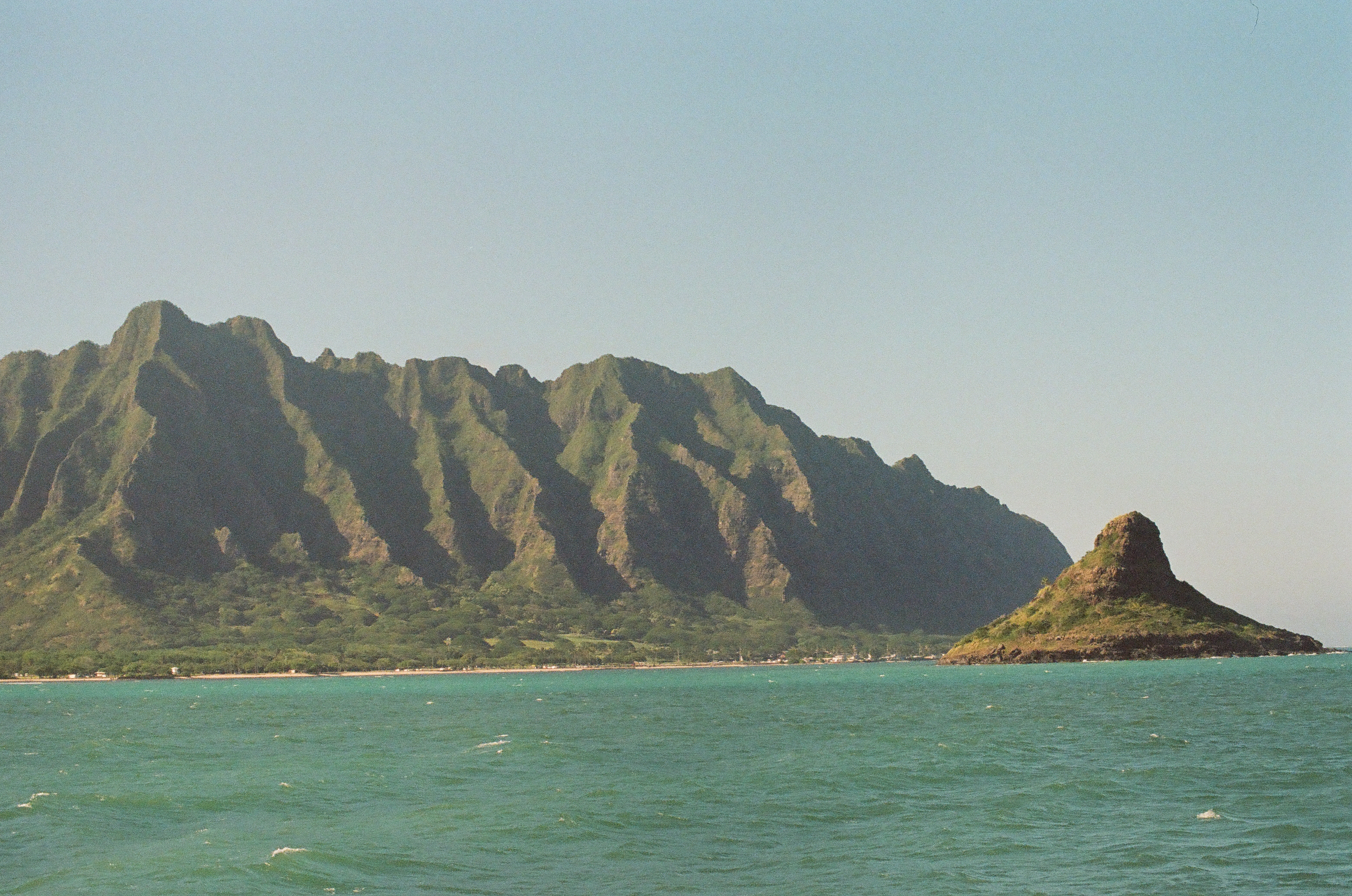 A jagged green mountain ridge fills the frame beside a rounded rocky peninsula, with sunlit ripples crossing the turquoise sea below a pale sky.