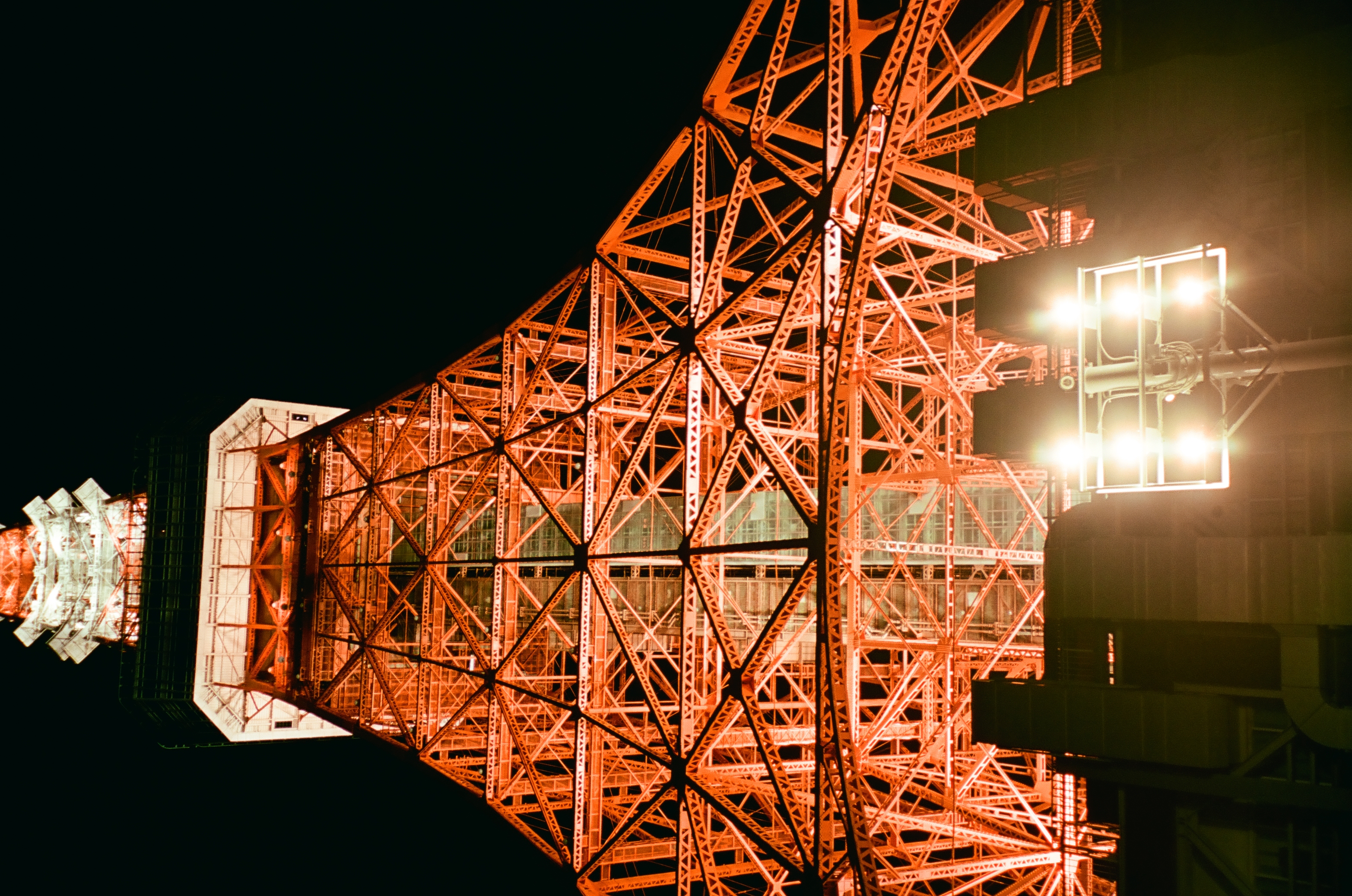 A multi-headed park floodlight blazes in the foreground below the full width of an orange lattice tower's base.