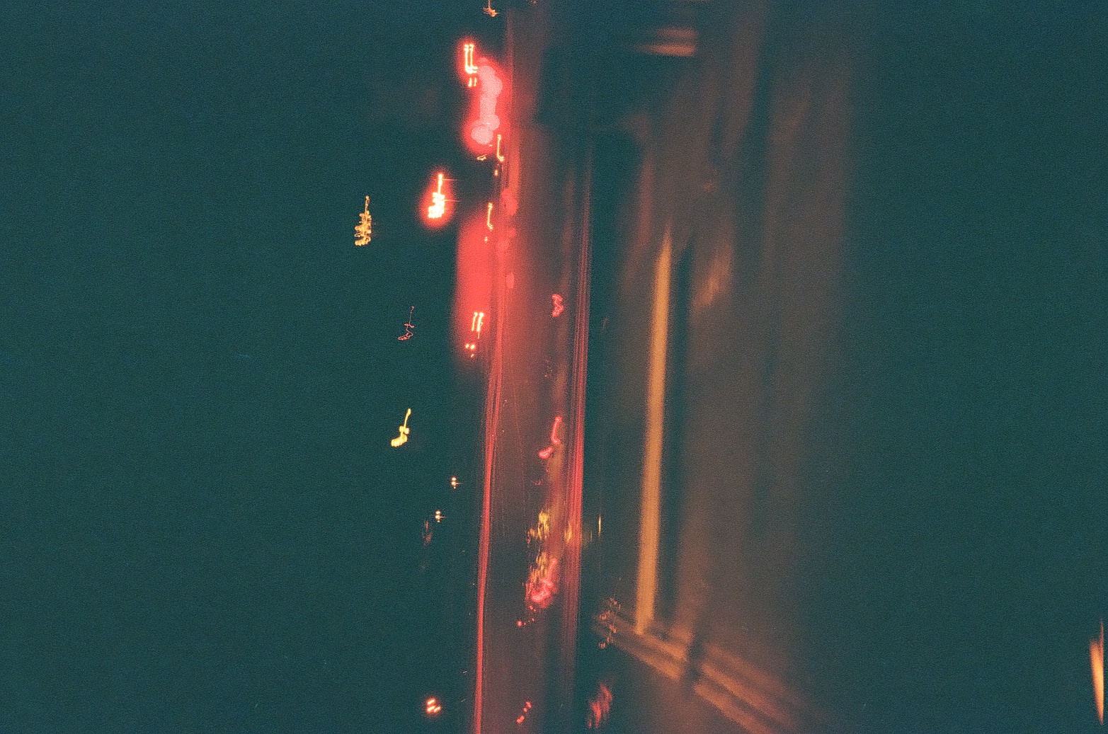 Blurred red and pale light trails streak diagonally along a dark road at night beside the shadowed edge of a building.