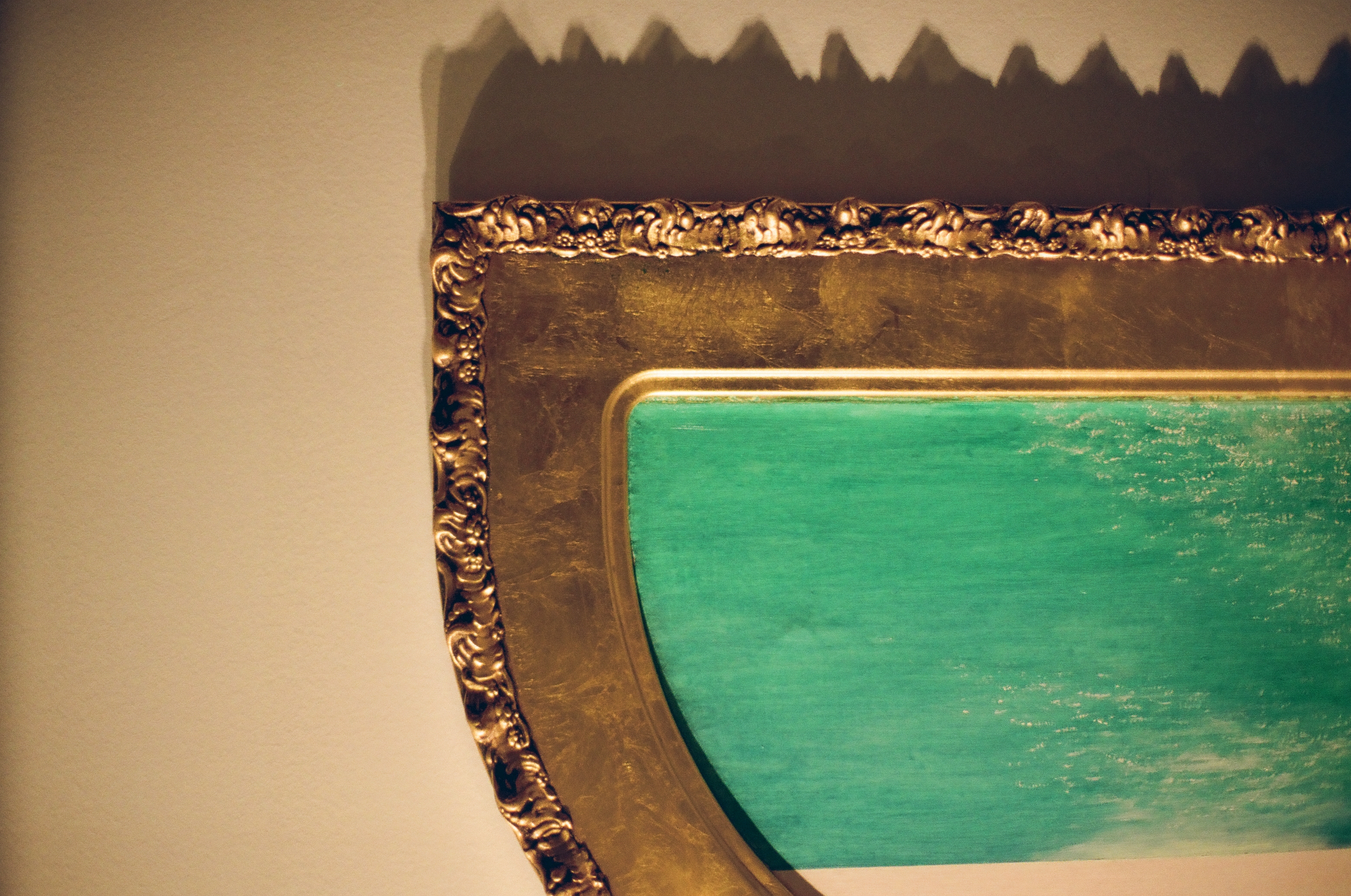 The lower right corner of an arched gold frame surrounds a field of green paint, casting a jagged sawtooth shadow onto the pale wall below.