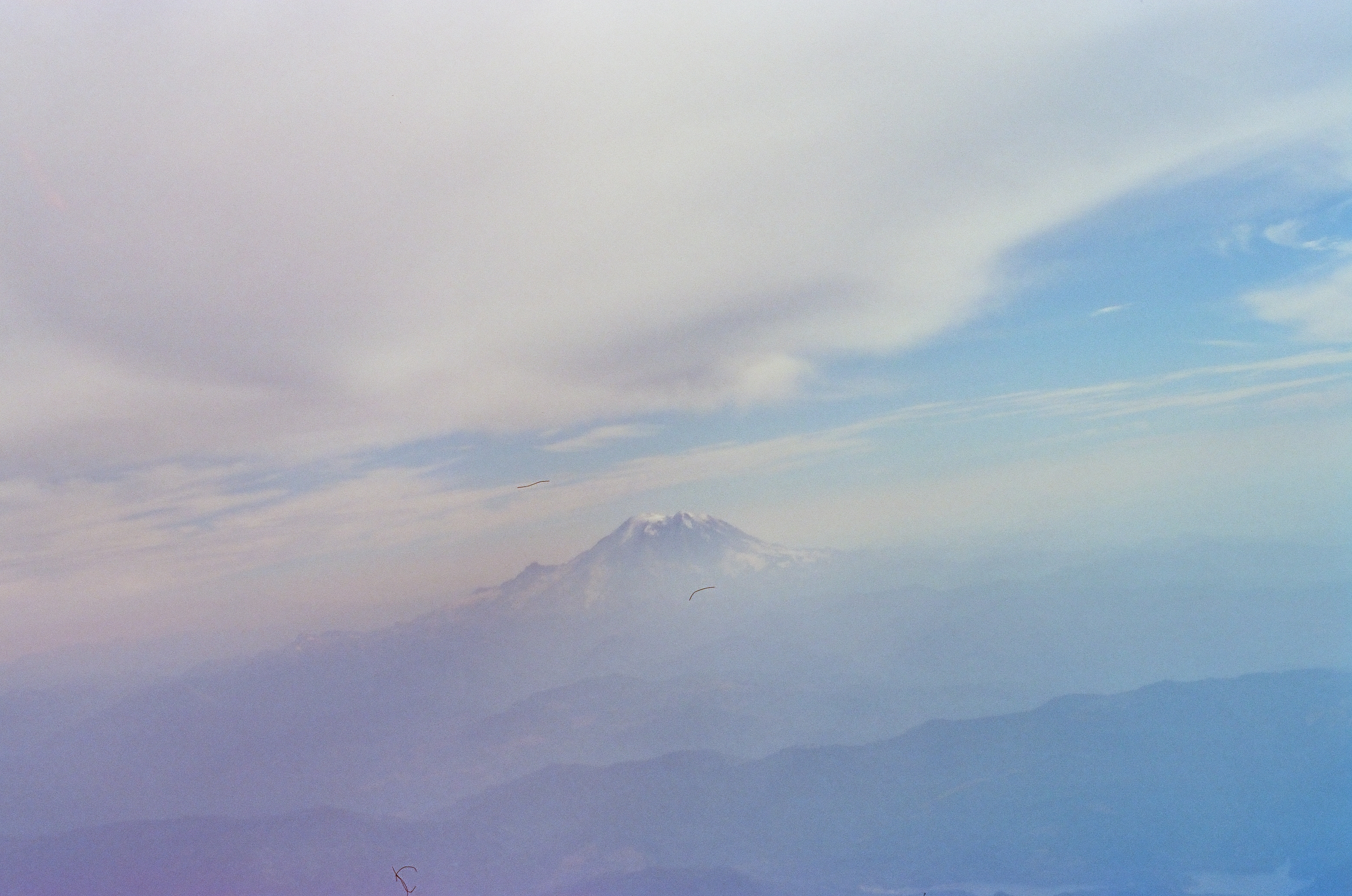 A snow-capped mountain peak appears faintly above layered blue ridgelines beneath a band of thick cloud, with two thin dark curved marks visible in the hazy sky.