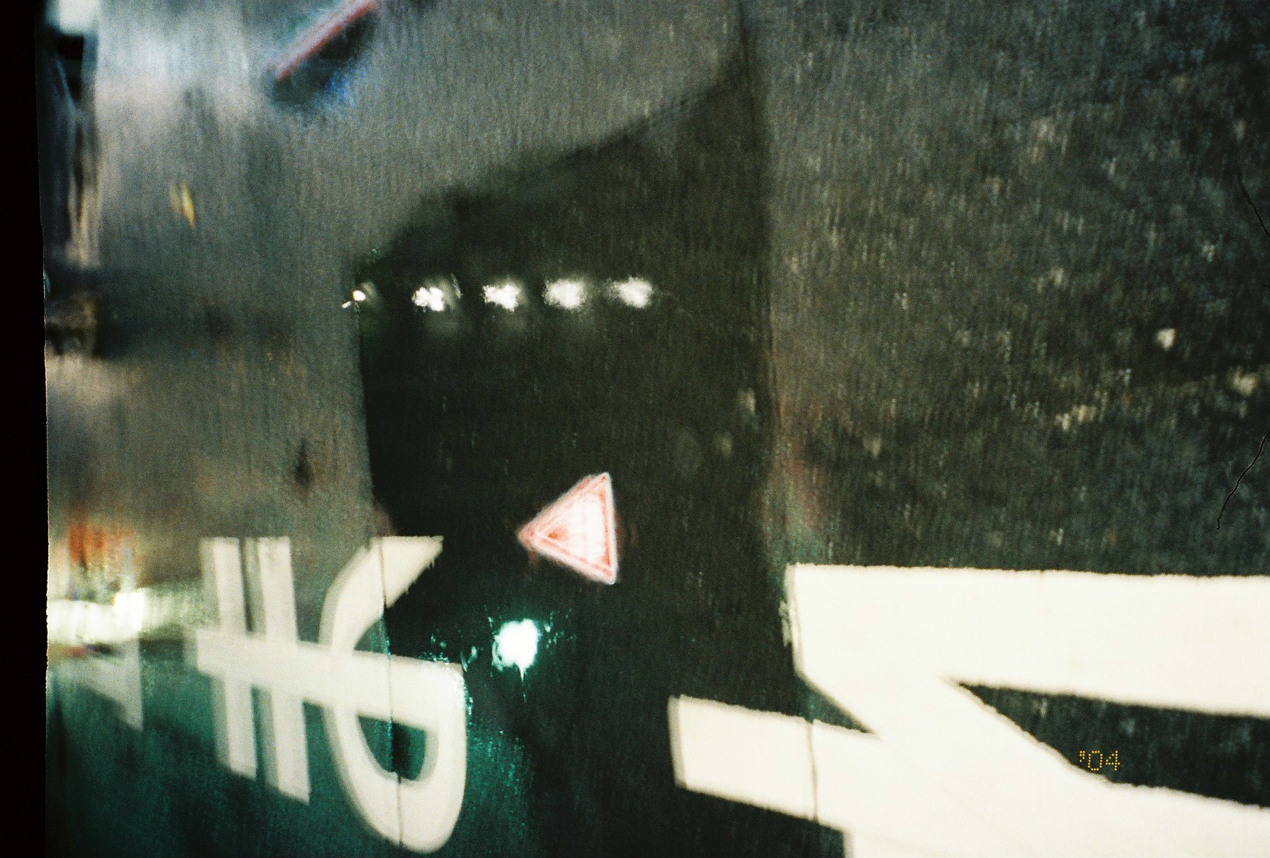 A rain-slicked street at night shows a white painted character and a red-and-white warning triangle floating among reflected lights on black pavement.