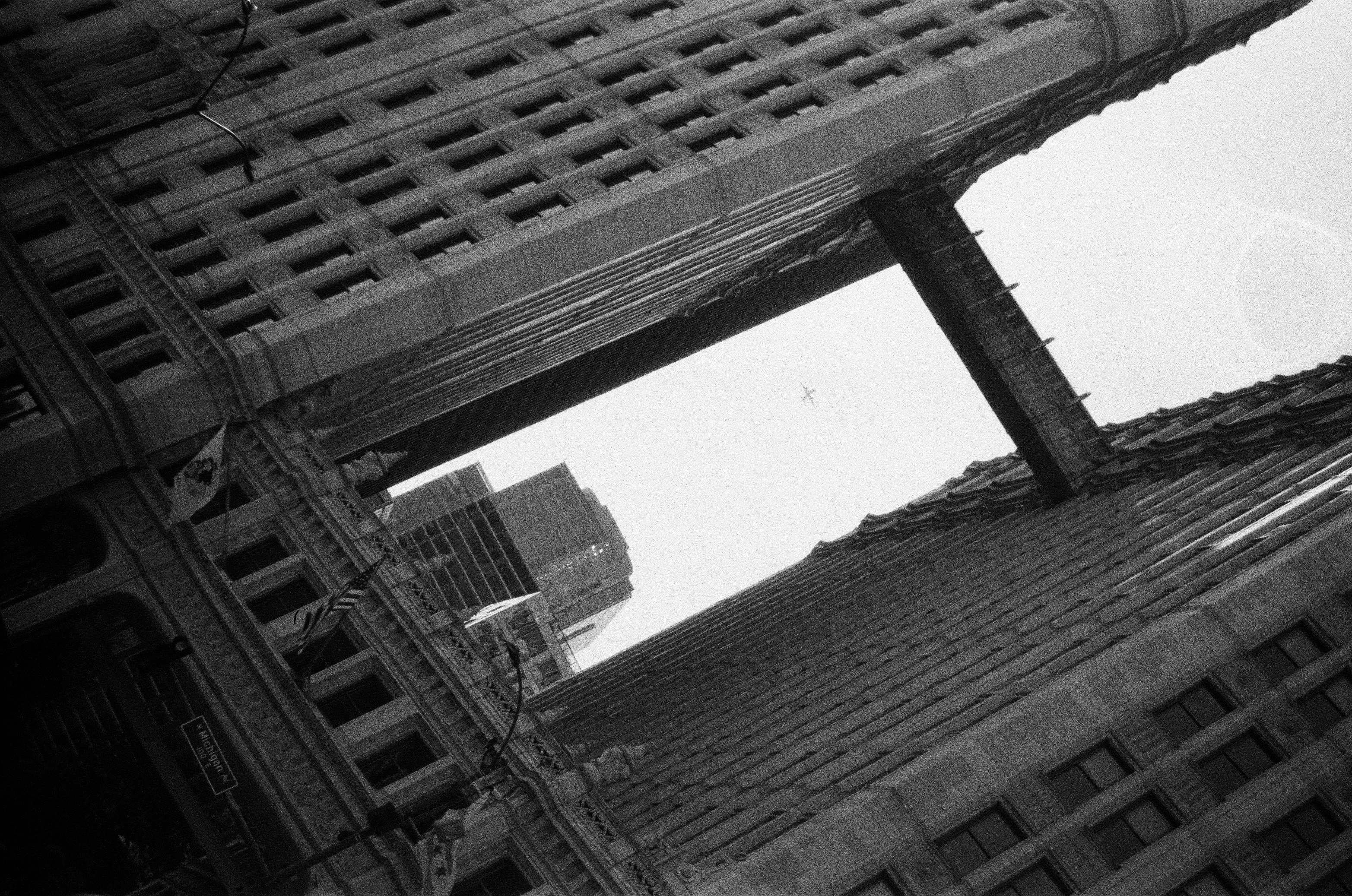A narrow vertical view looks upward between dark masonry towers joined by an elevated enclosed bridge.