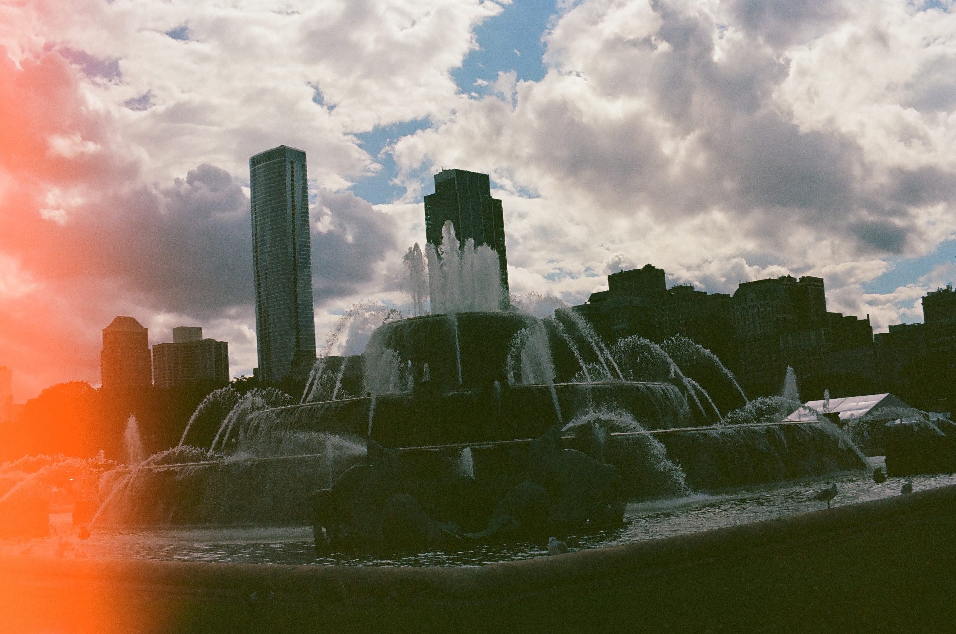 White fountain jets rise before a dark skyline as a red flare enters beneath brilliant summer clouds.
