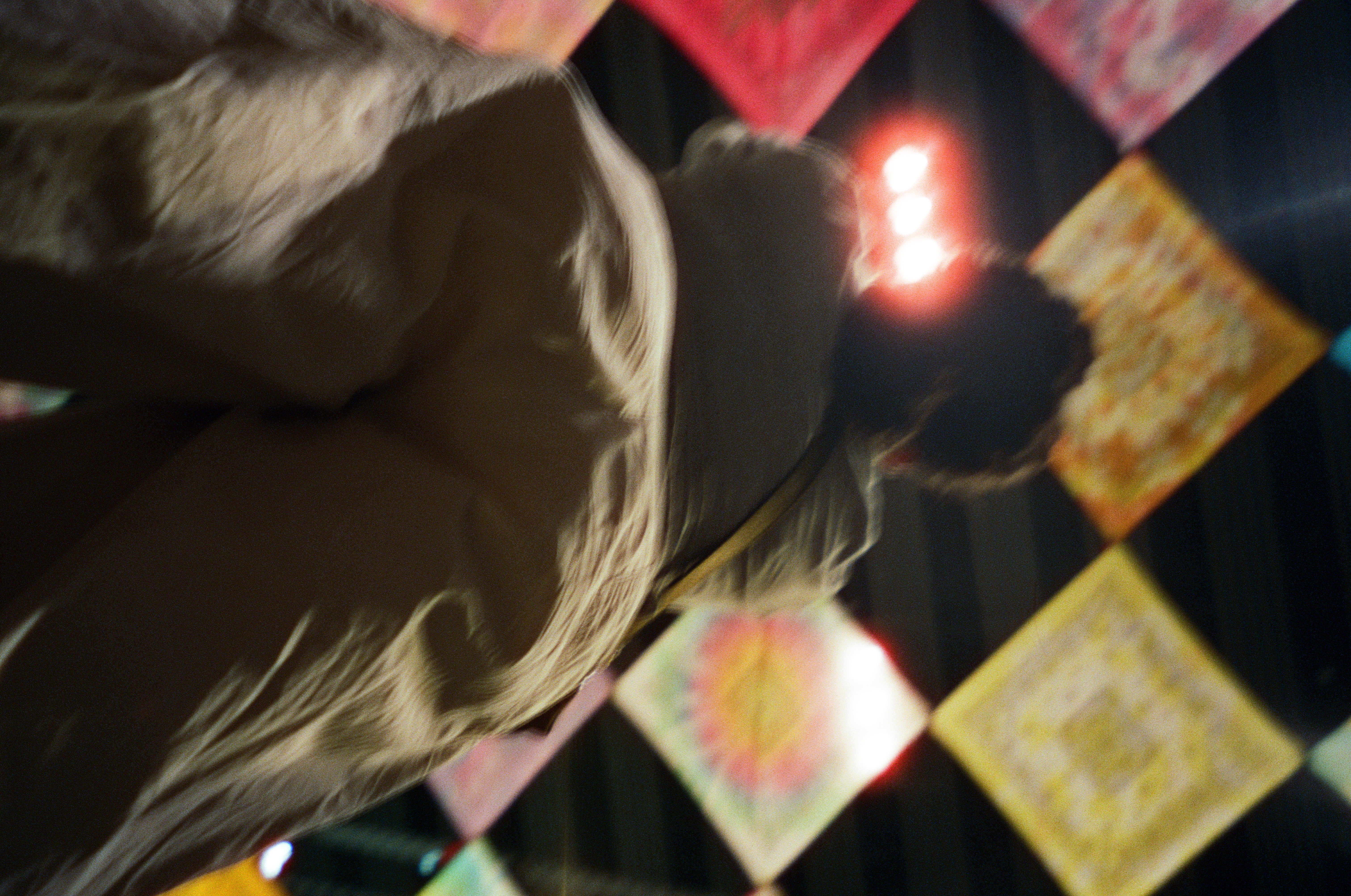 A motion-blurred person in a pale jacket is seen from below and behind, framed by diamond-hung tie-dye fabric panels overhead.