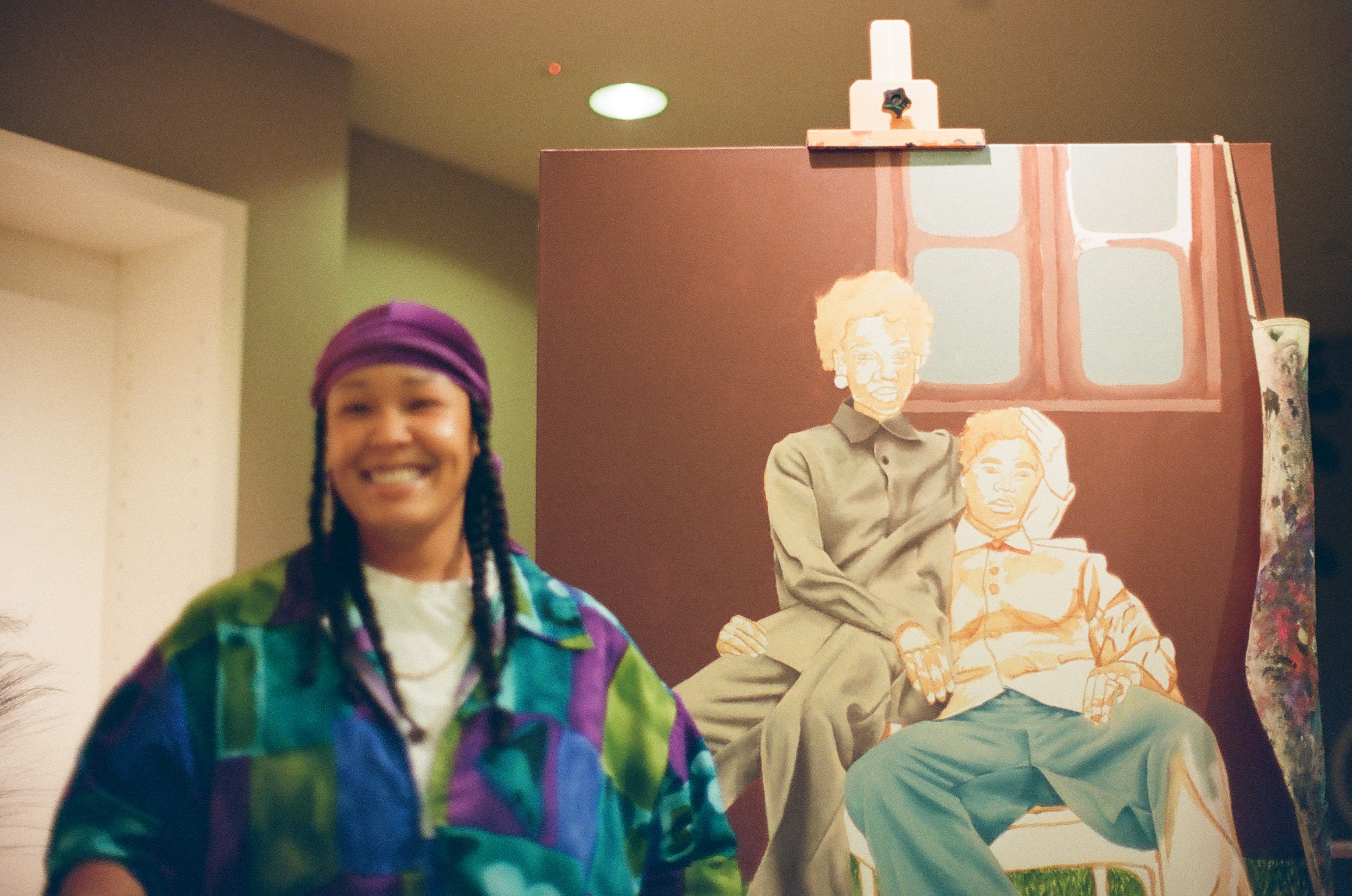 A smiling person appears softly blurred before a large easel painting depicting two seated figures.